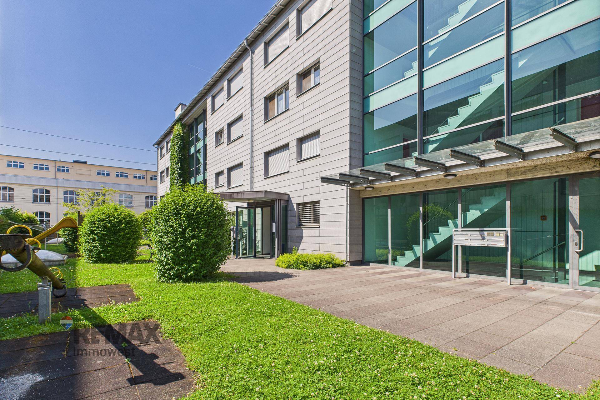 Modernes Bürogebäude mit Glasfassade und gepflegter Grünfläche im Eingangsbereich unter blauem Himmel.