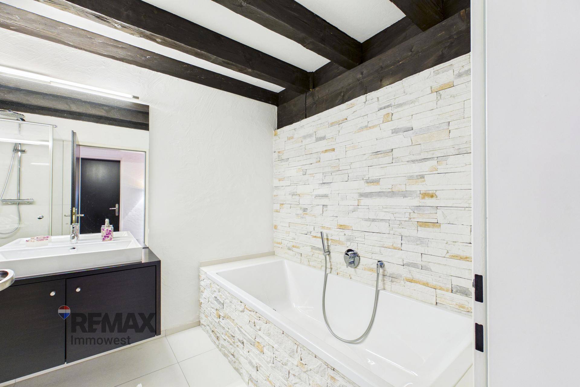 Modernes Badezimmer mit Badewanne, Steinwandakzent und sichtbaren Holzbalken an der Decke.