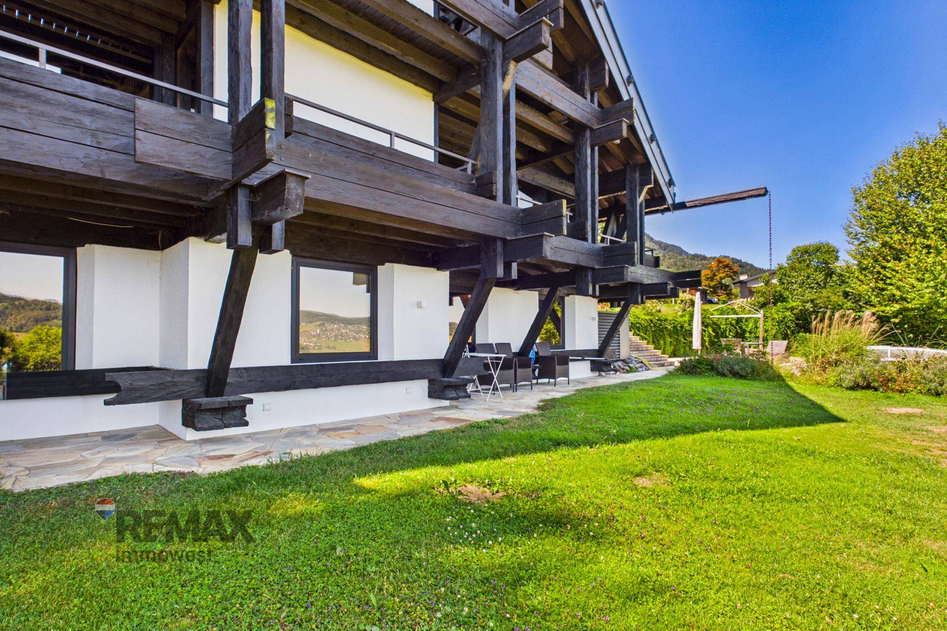 Rustikales Holzhaus mit weißer Fassade, umgeben von einem grünen Garten und Bergblick.