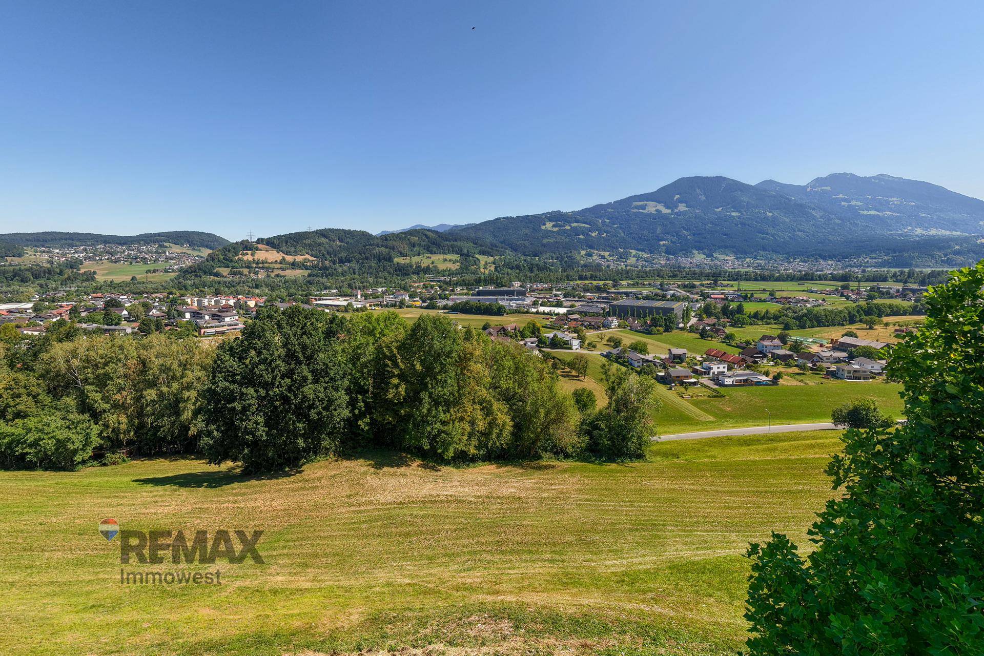 Weitläufiger Blick über die grüne Landschaft mit Bergen und einer Siedlung in der Ferne.