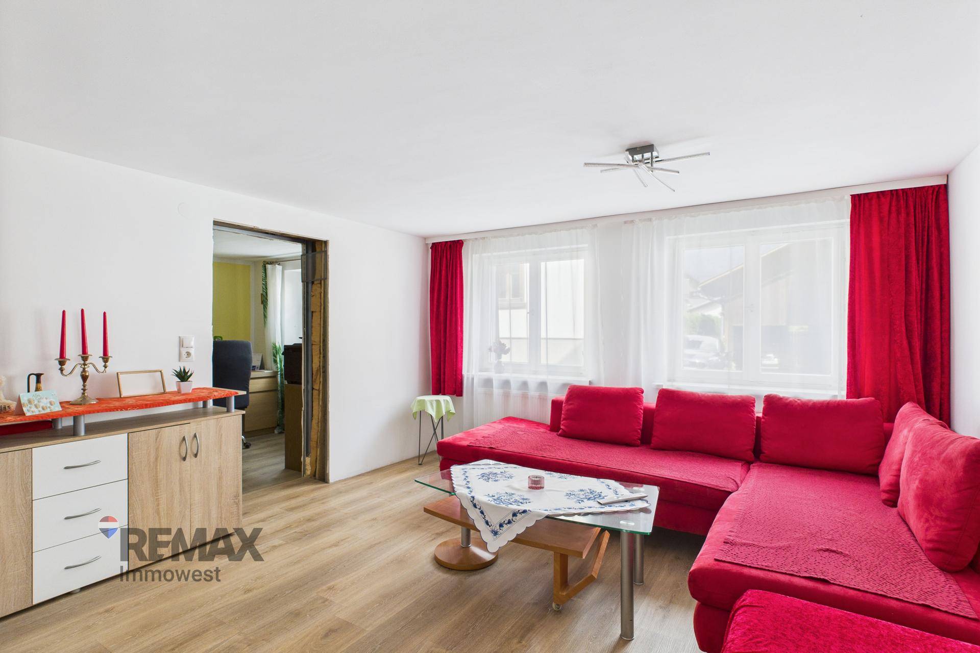 Helles Wohnzimmer mit rotem Sofa, großen Fenstern und Zugang zu einem weiteren Raum.