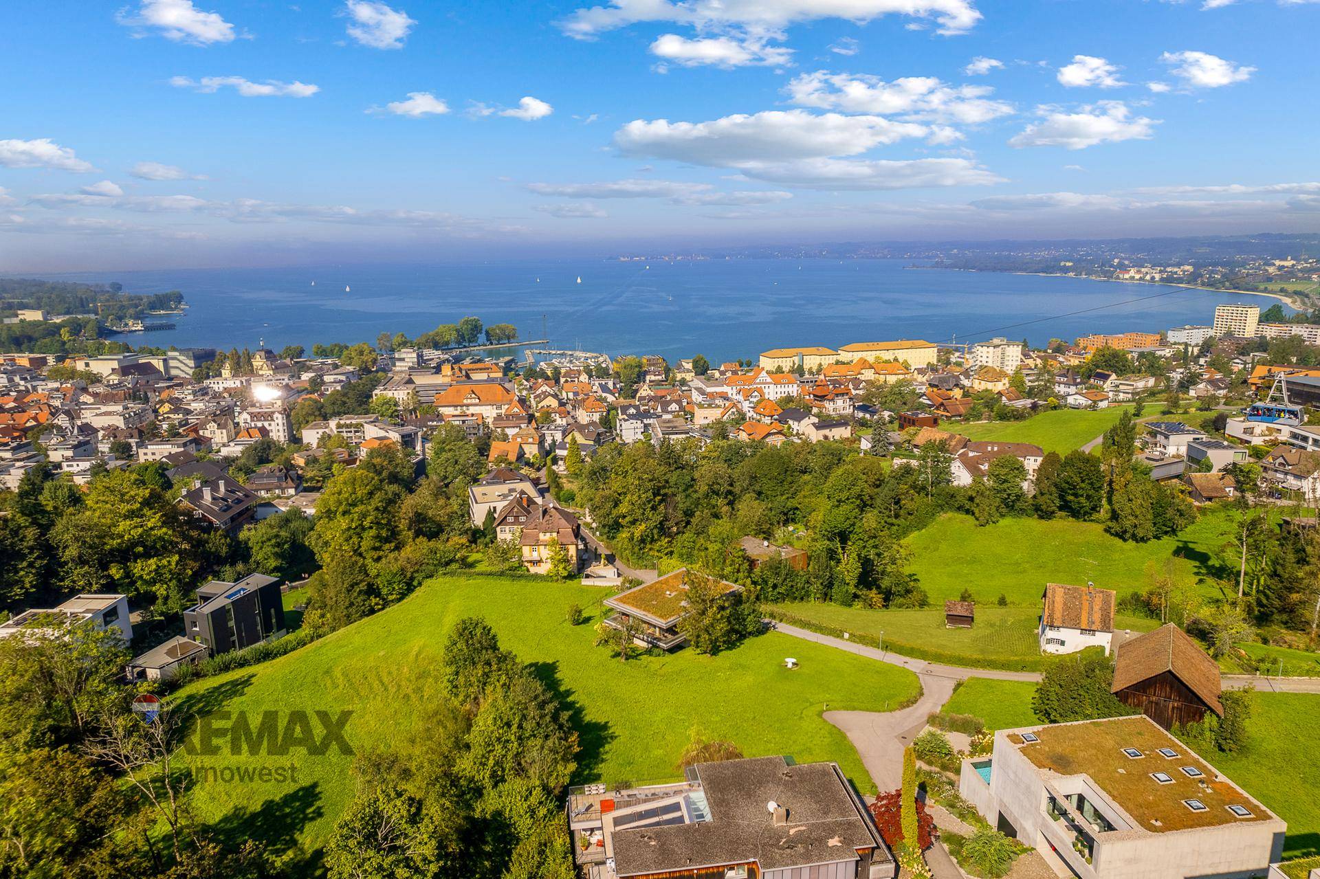 Weitläufige Luftaufnahme einer Stadt am Seeufer, umgeben von grünen Hügeln unter einem blauen Himmel.