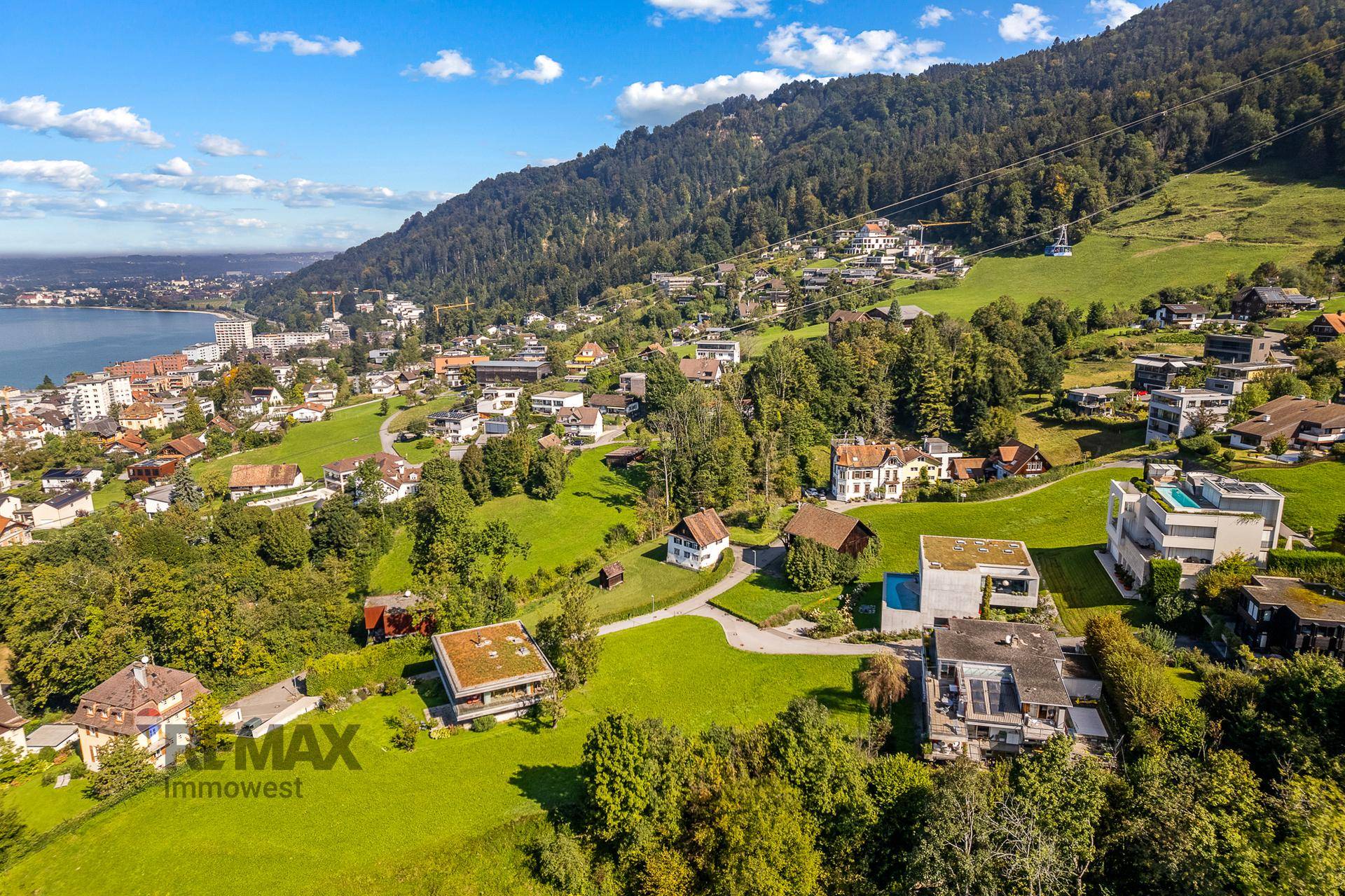 Malerische Luftaufnahme einer Stadt am See, umgeben von Bergen mit einer Seilbahn und üppiger Vegetation.