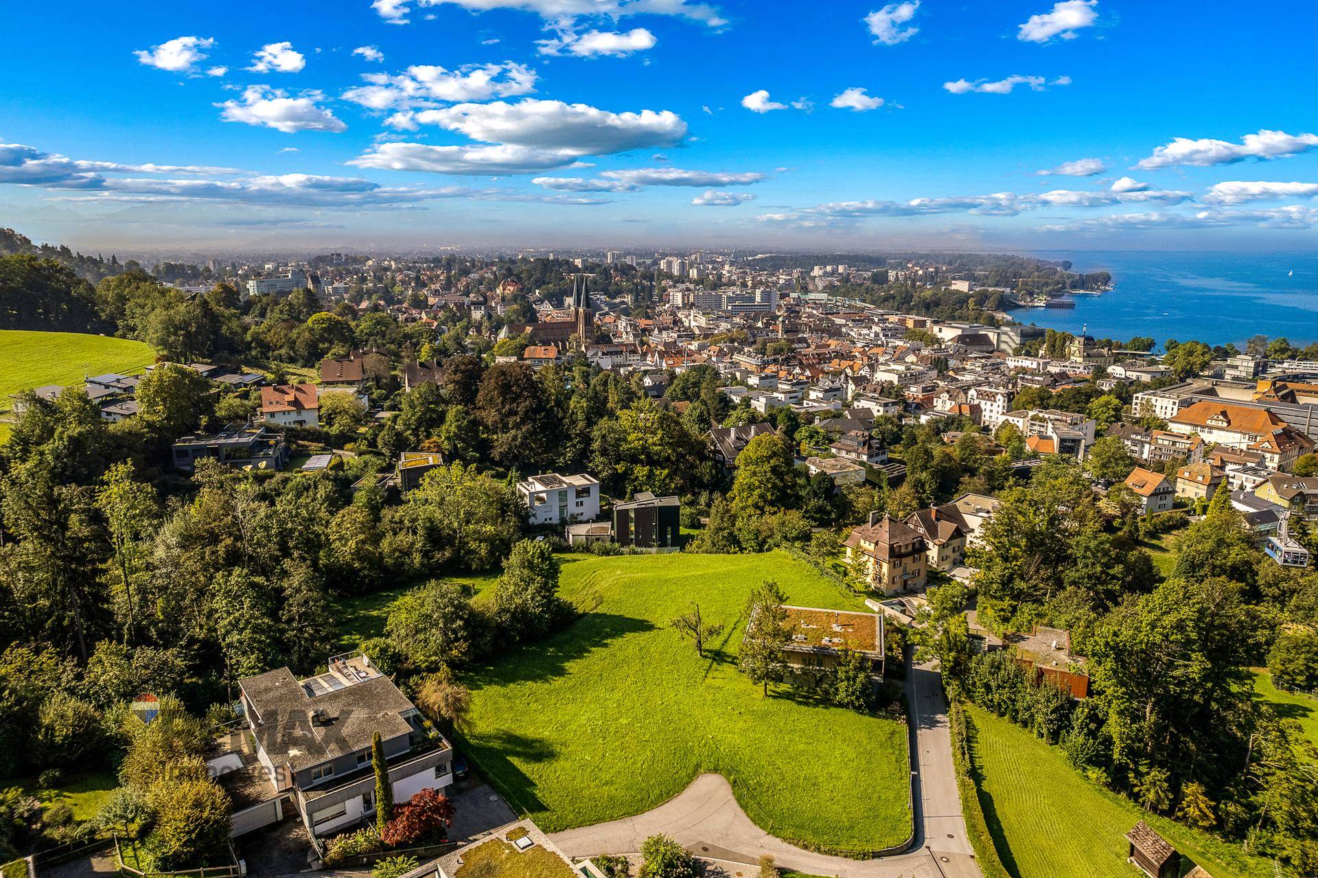 Panoramablick aus der Vogelperspektive auf eine Stadt, einen See und die umliegende grüne Hügellandschaft.