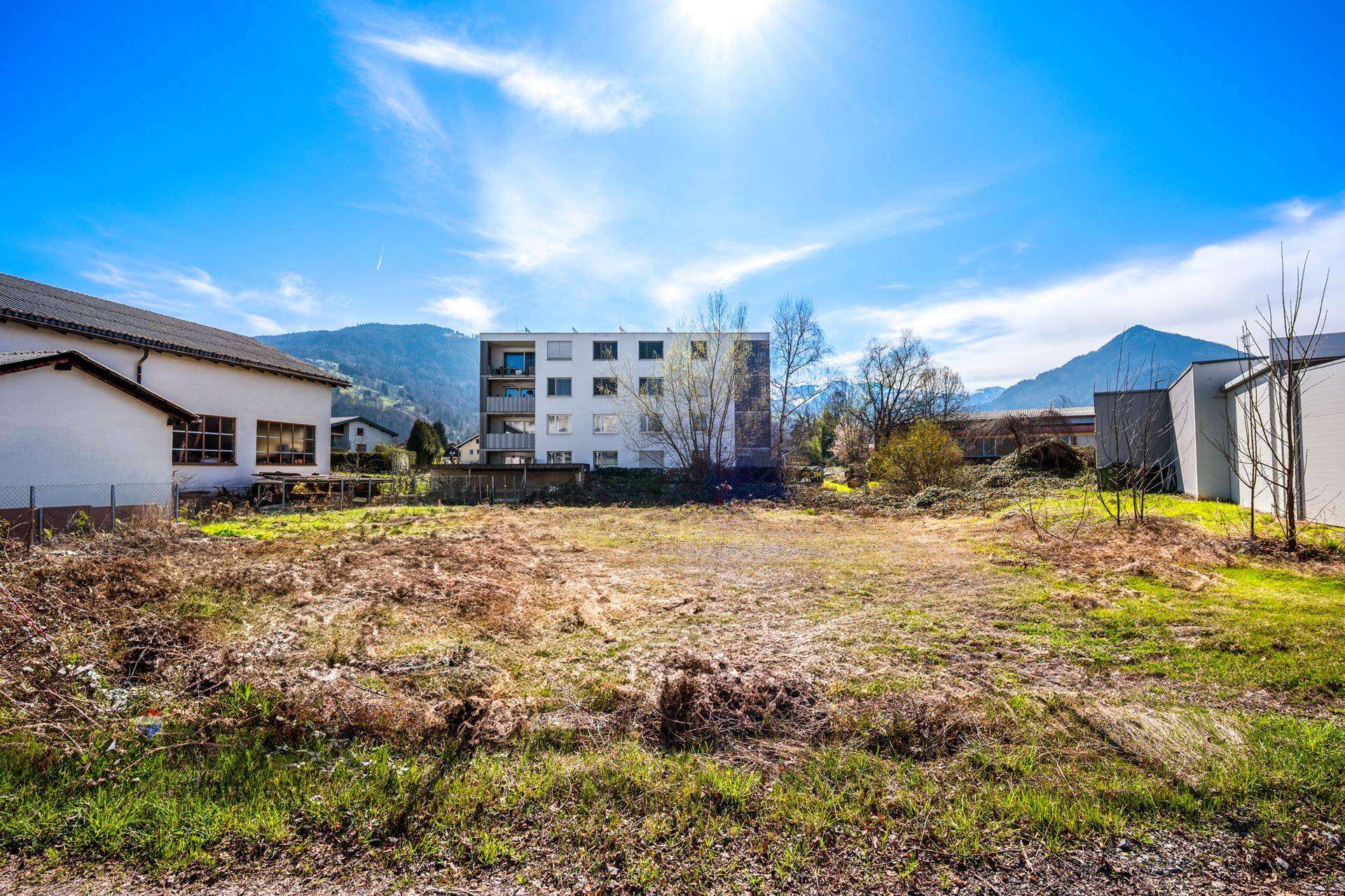 Sonniger Blick auf ein freies Baugrundstück mit angrenzenden Gebäuden und einem weiten Bergpanorama.