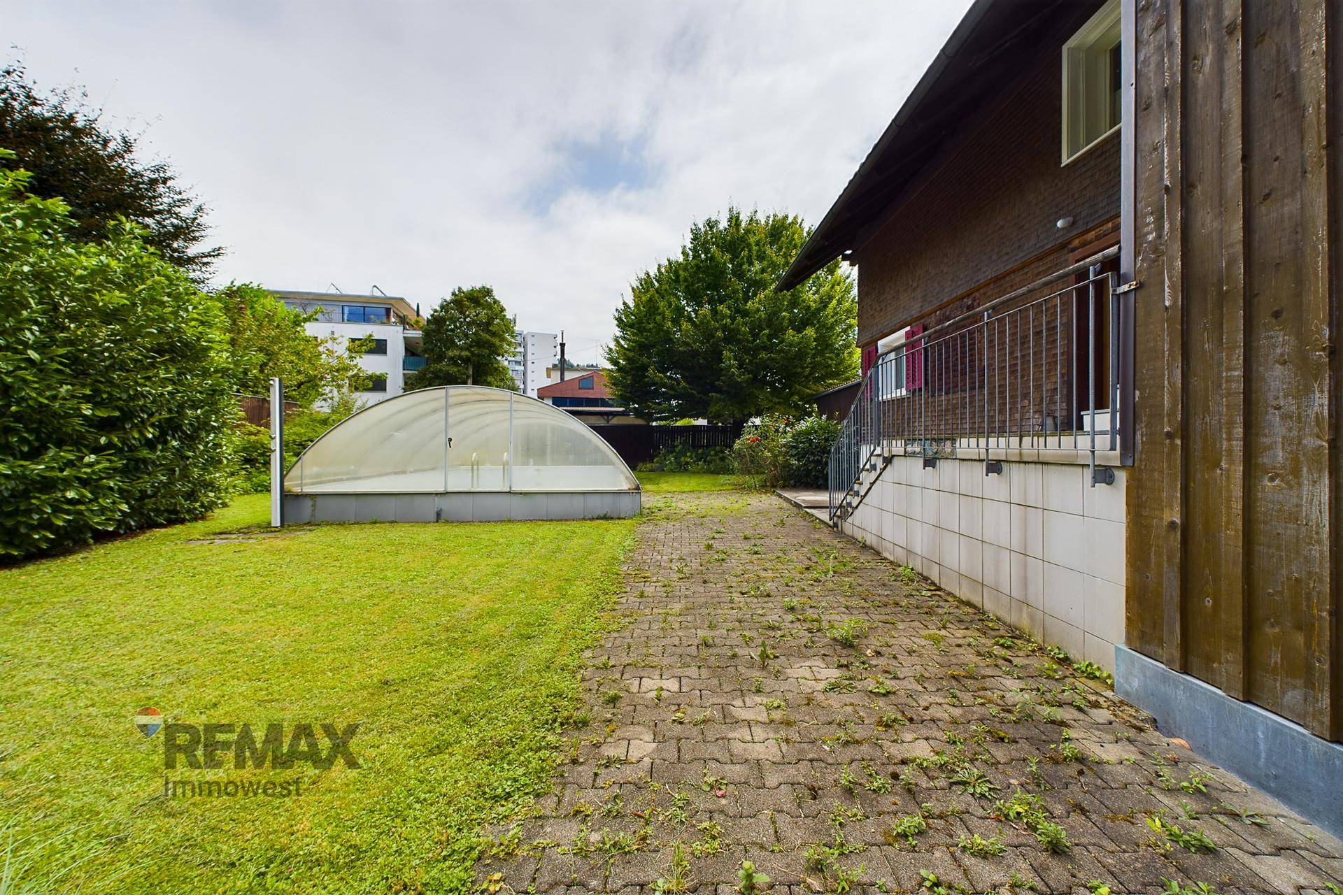 Seitenansicht eines Hauses mit gepflastertem Weg, Rasenfläche und einem Gewächshaus im Garten.