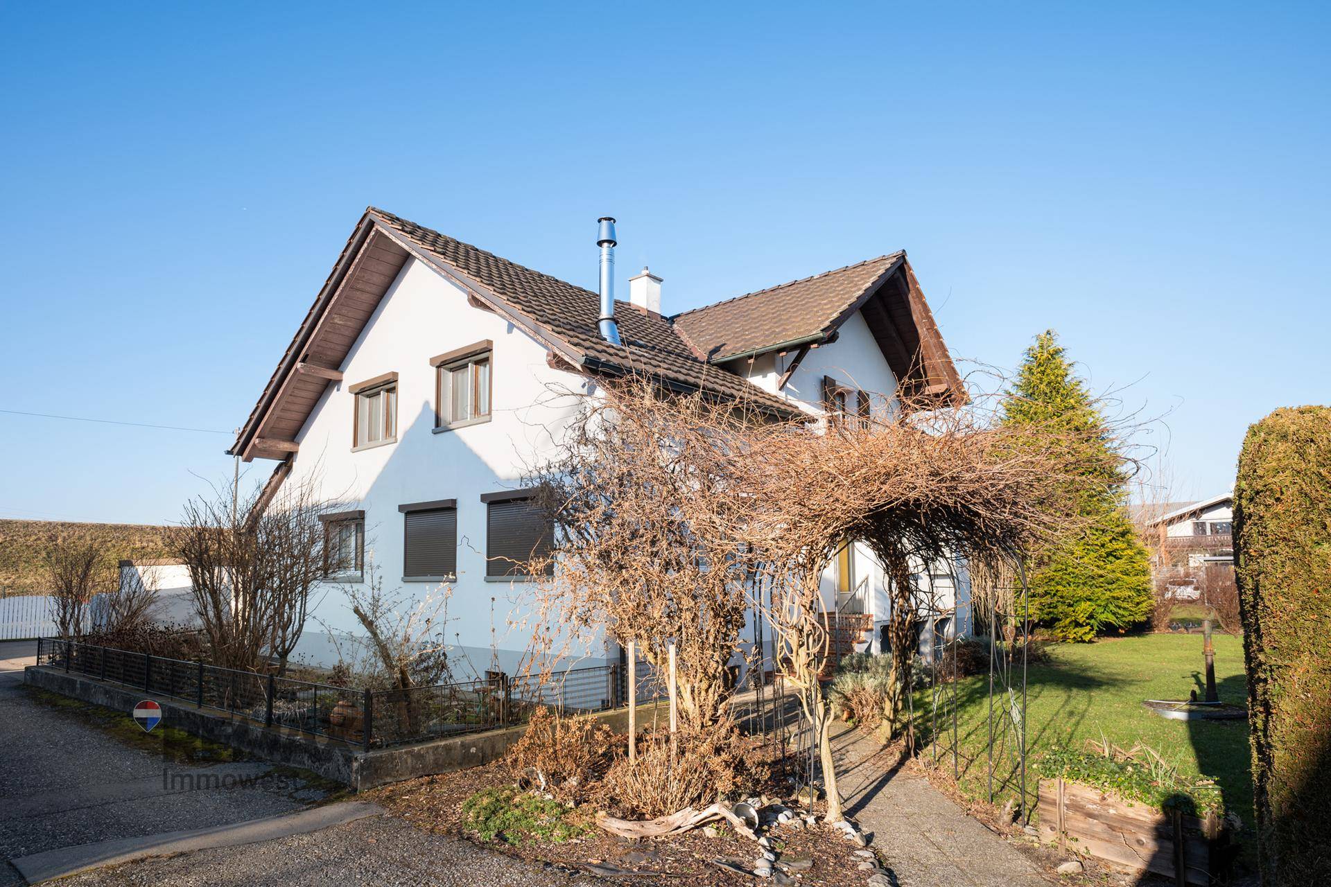 Weißes Einfamilienhaus mit braunem Dach, umgeben von einem kleinen Garten und einer Pergola.