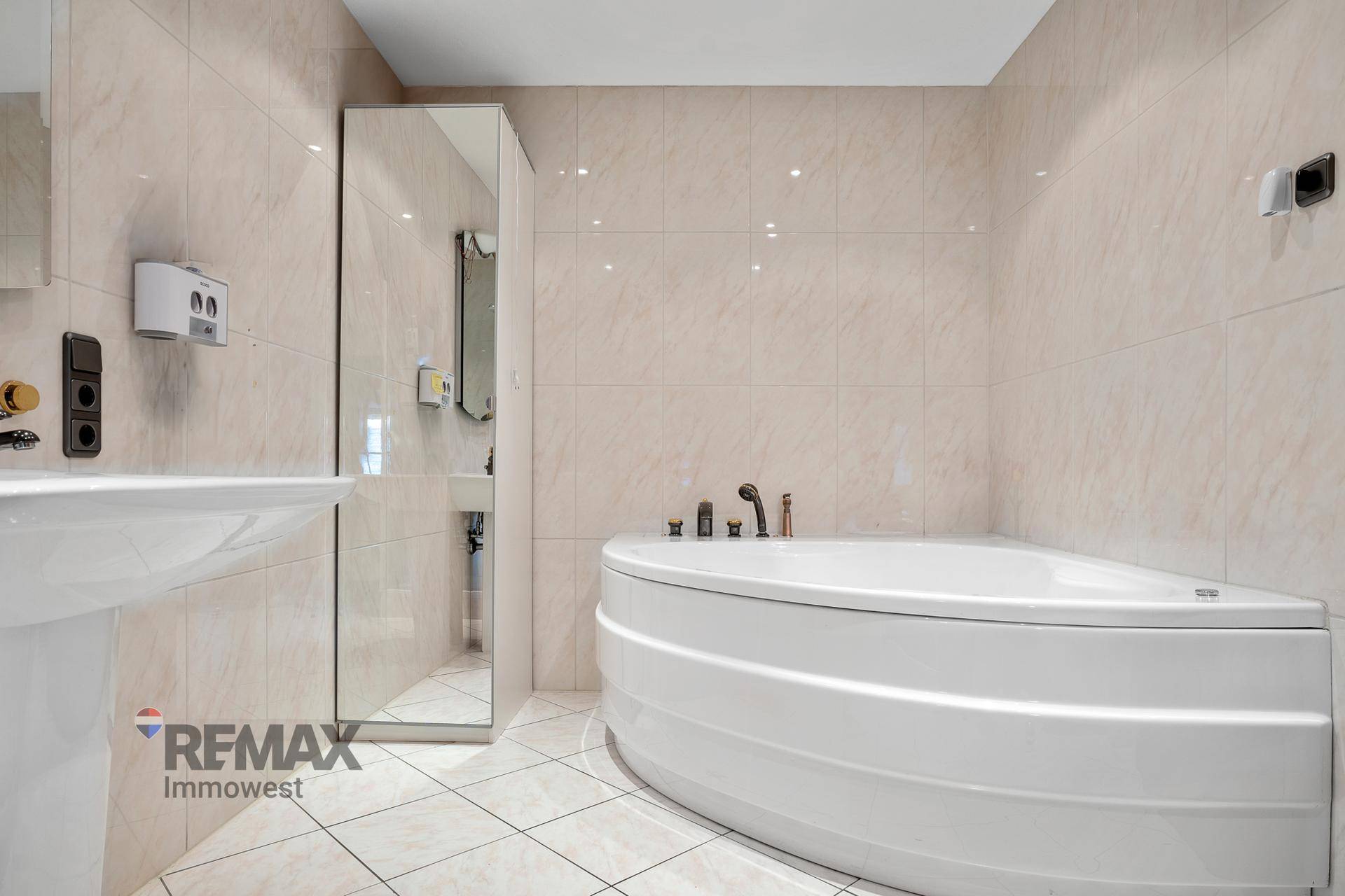 Badezimmer mit beigefarbenen Fliesen, einer Eckbadewanne und einem Standwaschbecken.