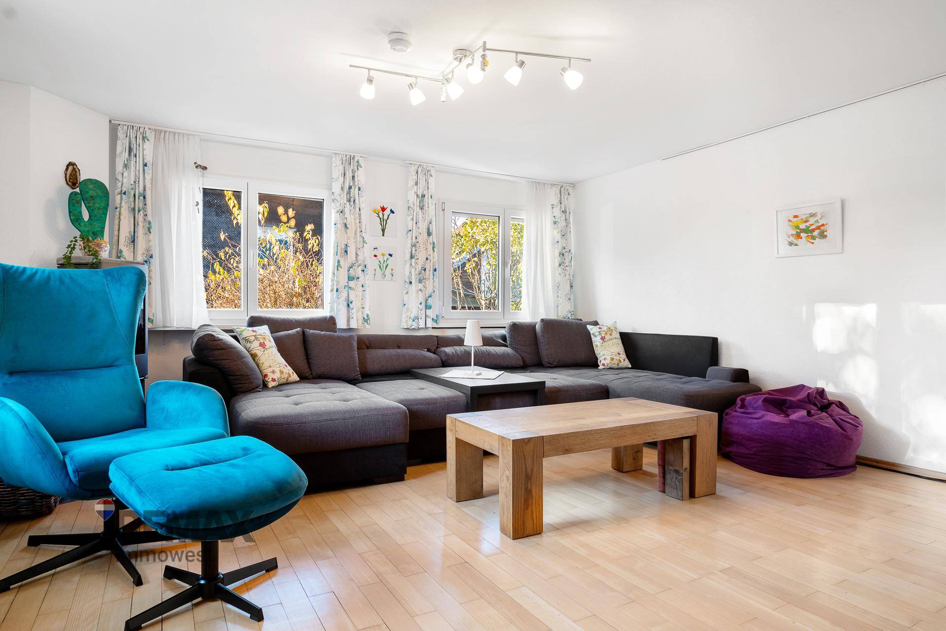 Helles Wohnzimmer mit großem Ecksofa, Holztisch und einem blauen Sessel, Parkettboden.