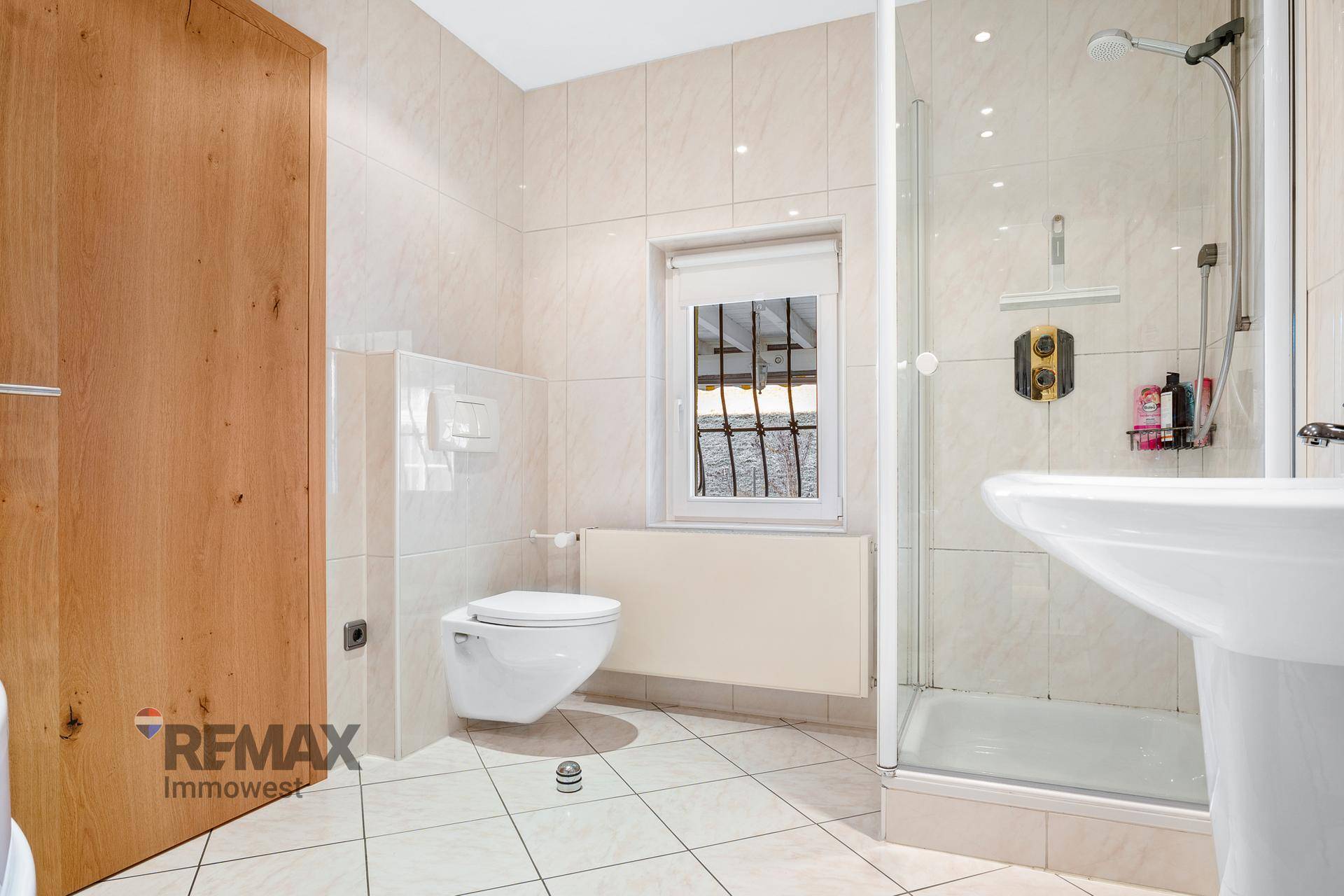Badezimmer mit beigefarbenen Fliesen, wandhängendem WC, Duschkabine und Fenster.