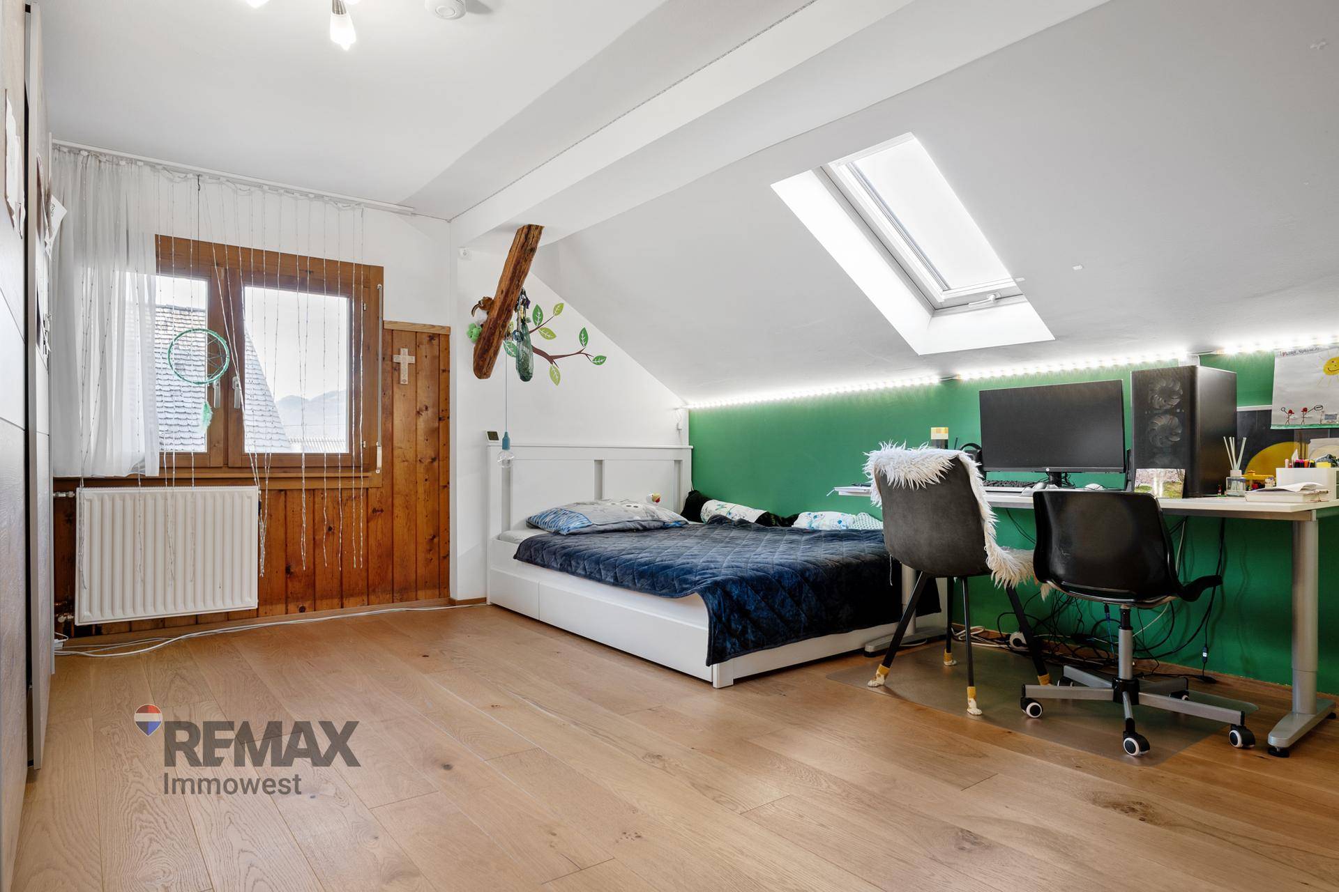 Schlafzimmer mit Bett, Schreibtisch, grüner Akzentwand, Dachfenster und Holzboden.
