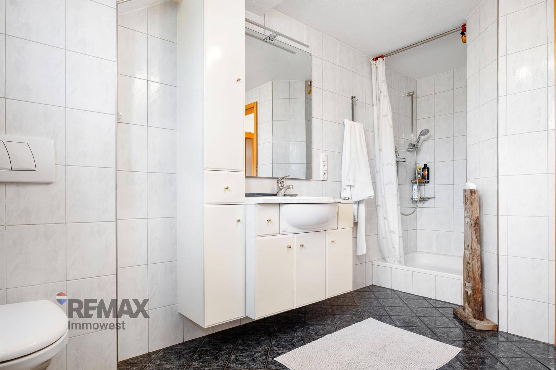 Badezimmer mit weißen Wandfliesen, dunklen Bodenfliesen, Dusche und Waschtisch.