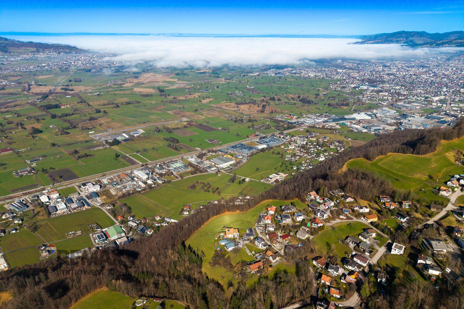 Ausgedehnte Luftaufnahme einer weiten Landschaft mit Feldern, Wäldern und einer Siedlung unter einer Nebeldecke.