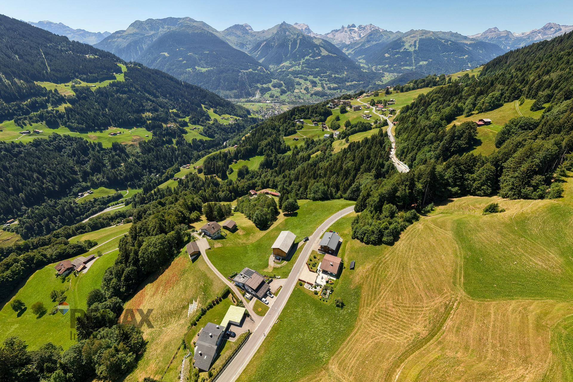 Panoramablick auf ein Tal mit verstreuten Häusern, grünen Hängen und majestätischen Bergen im Hintergrund.