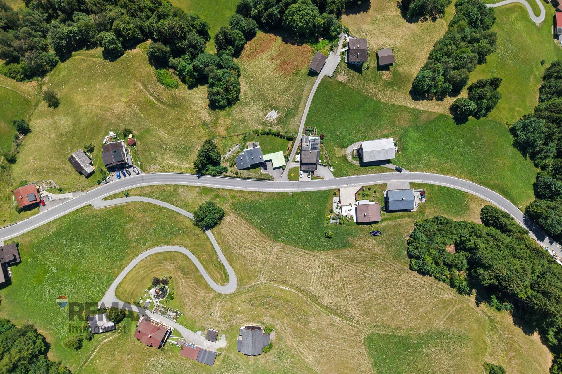 Weitläufige Luftaufnahme einer ländlichen Siedlung mit Straßen und Häusern in einer grünen Berglandschaft.