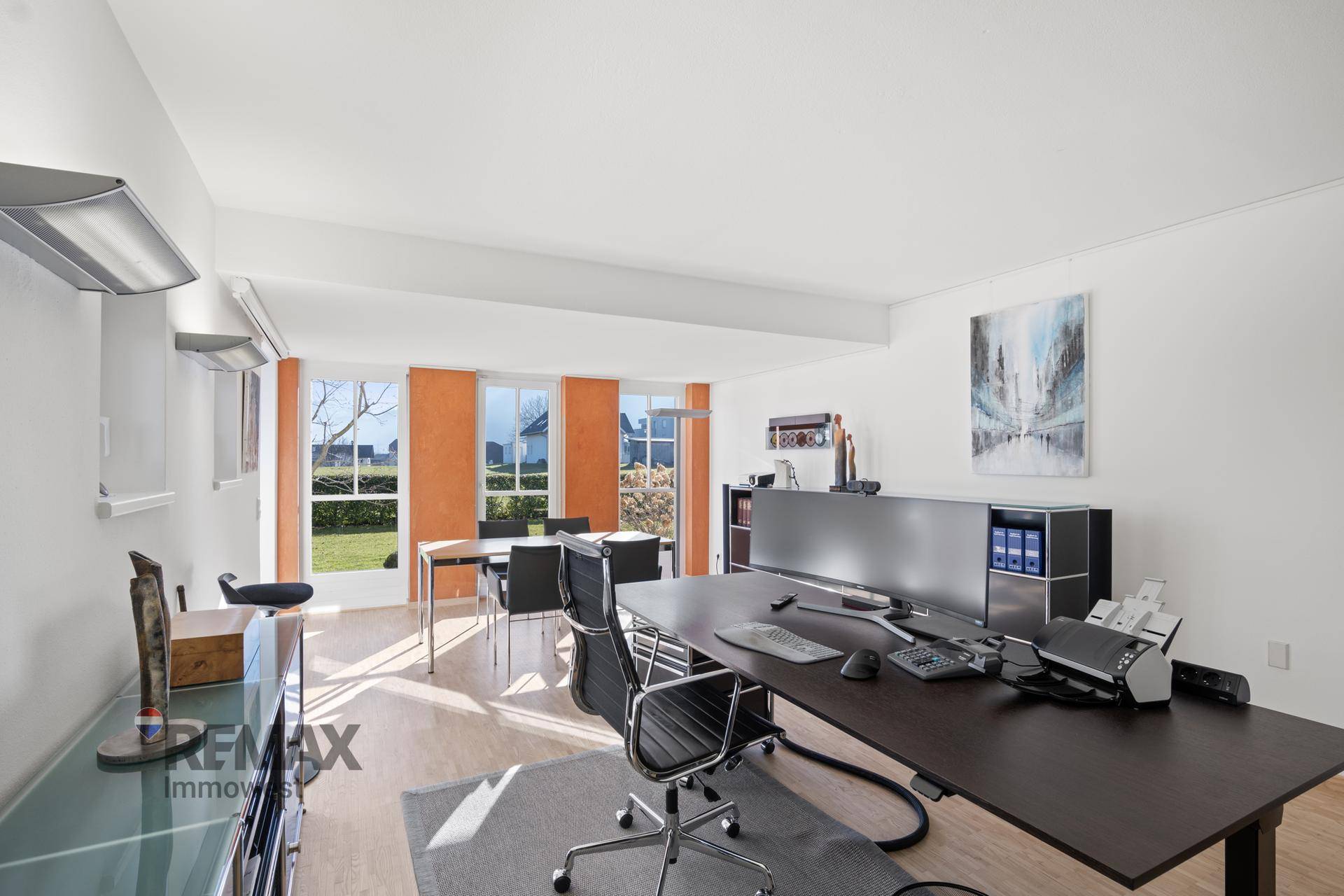 Helles Büro mit großem Schreibtisch, Holzboden und Blick ins Grüne durch mehrere Fenster.