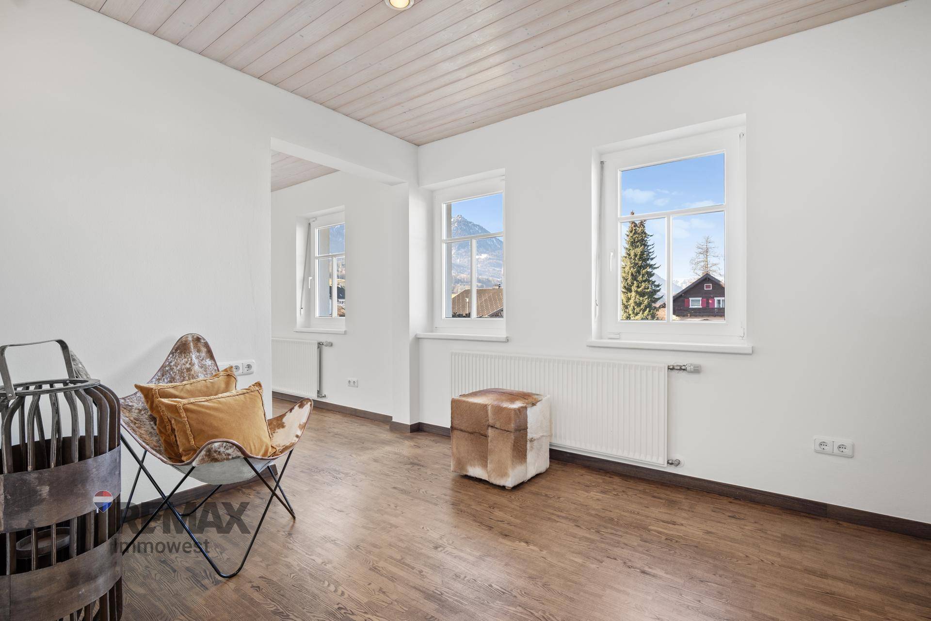 Heller Raum mit Holzboden, Fensterblick auf die Berge und dekorativen Sitzmöbeln.