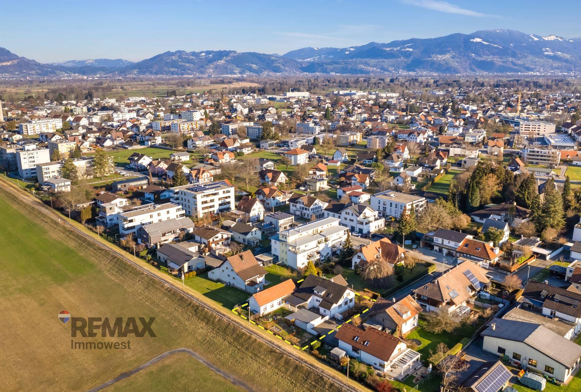 Weitläufige Luftaufnahme einer Wohnsiedlung mit Feldern und Bergen am Horizont unter blauem Himmel.