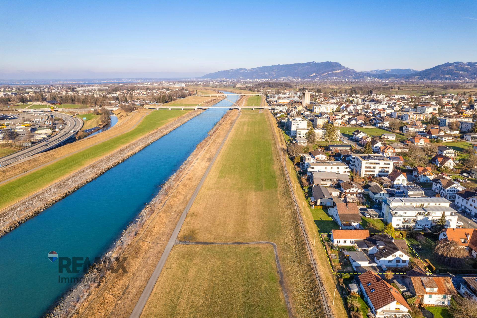 Luftaufnahme mit Fluss, Wohngebiet und Bergkette im Hintergrund unter blauem Himmel und Sonnenschein.