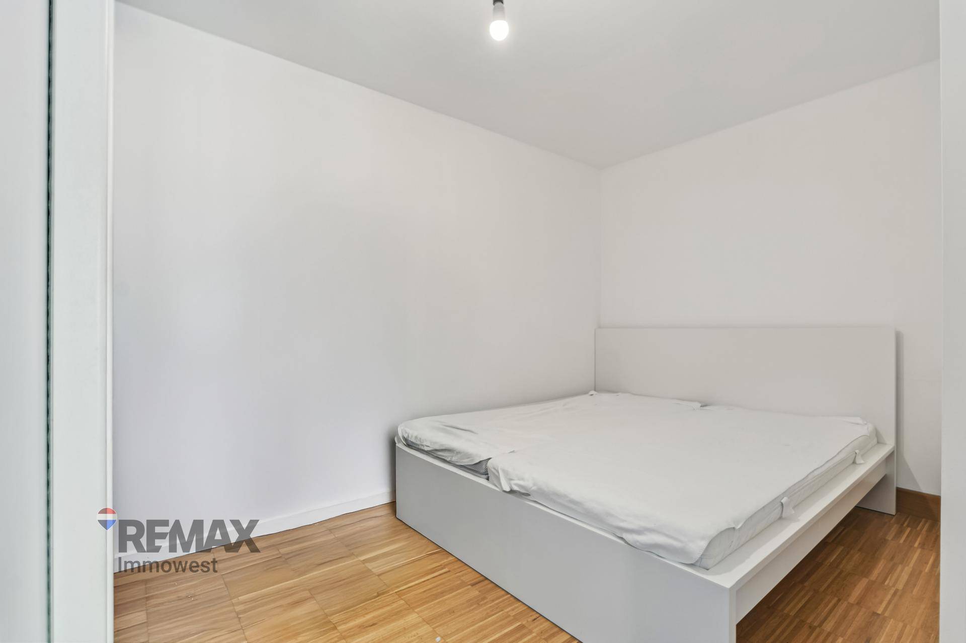 Schlichtes Schlafzimmer mit weißem Bettrahmen und hellen Wänden, ausgestattet mit einem warmen Holzfußboden.