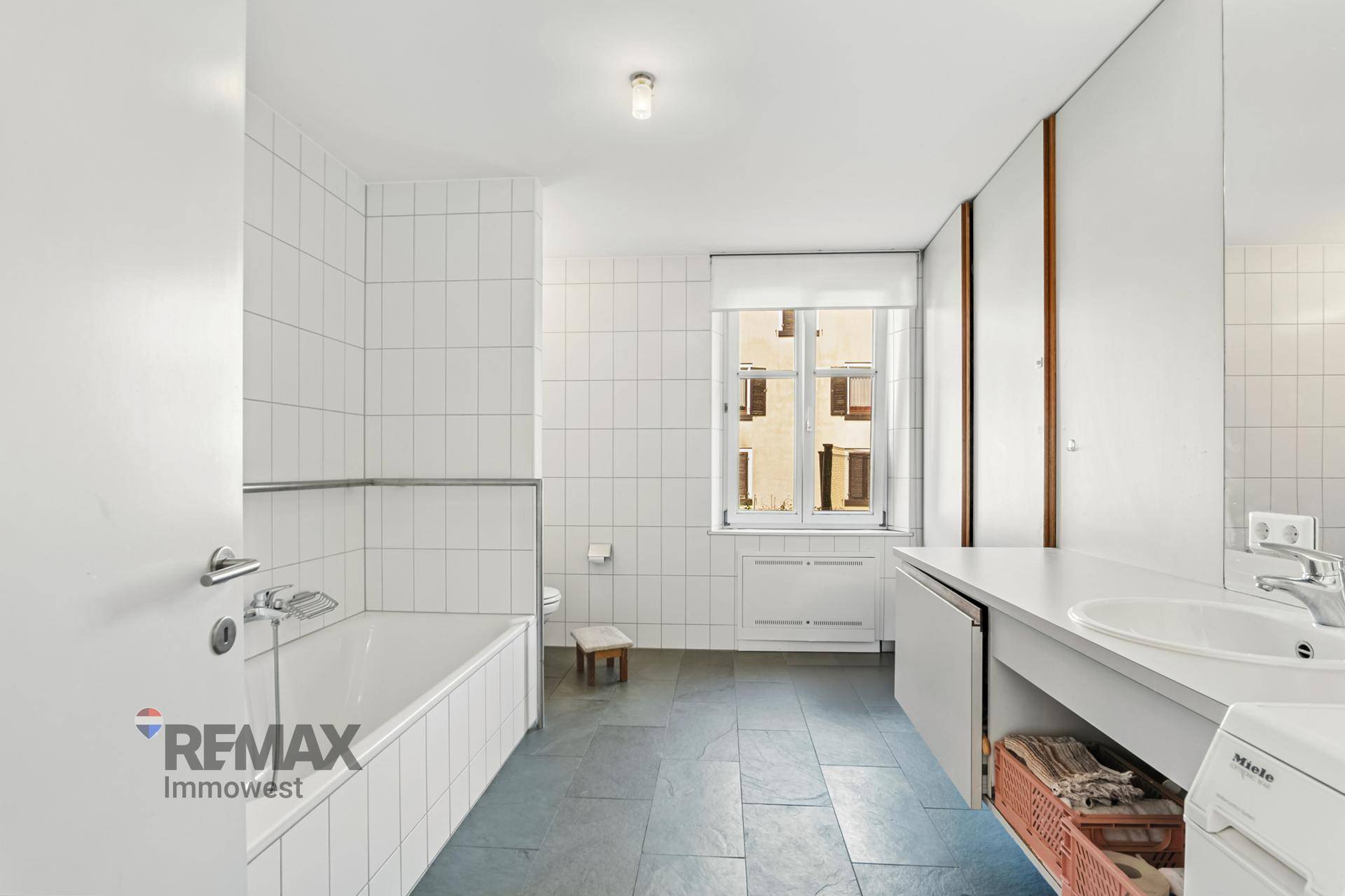 Helles Badezimmer mit Badewanne, Waschbecken und Fenster, ausgestattet mit weißen Fliesen und dunklem Boden.