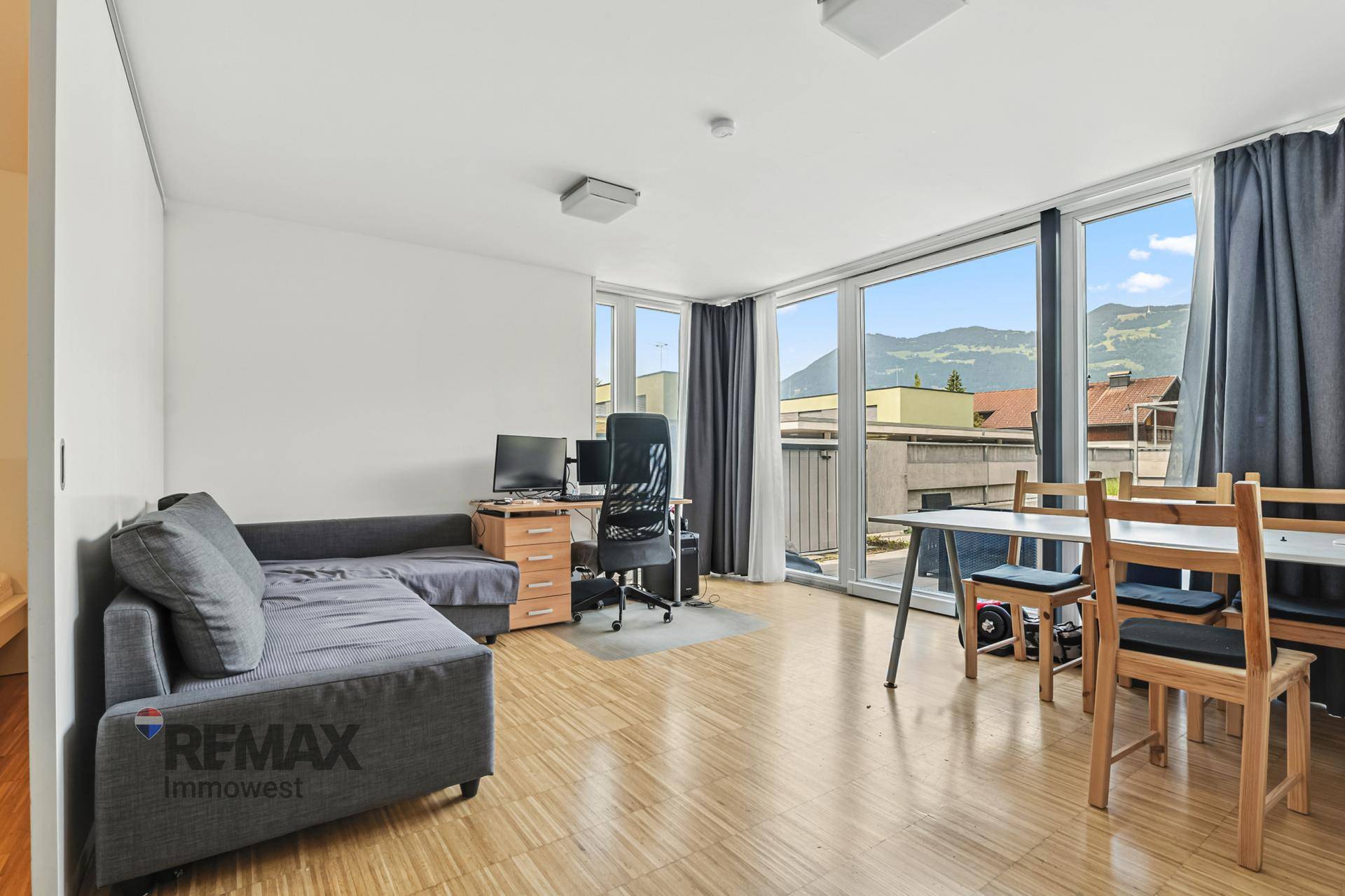 Geräumiges Wohnzimmer mit Sofa, Arbeitsbereich und Essplatz, großen Fenstern und Bergblick.