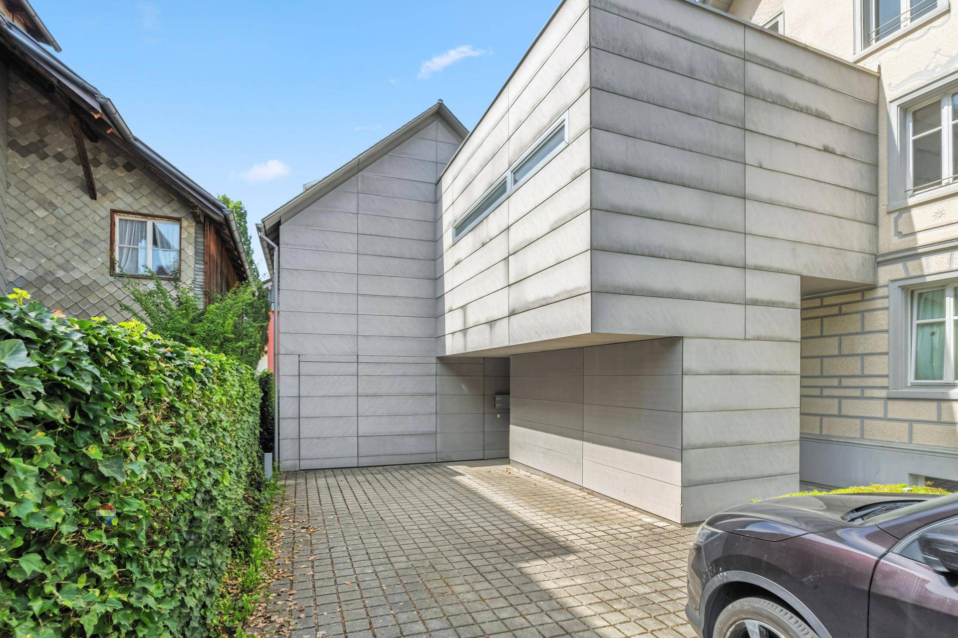 Moderne Gebäudefassade mit grauen Paneelen und einer Einfahrt, angrenzend an ein traditionelles Wohnhaus.