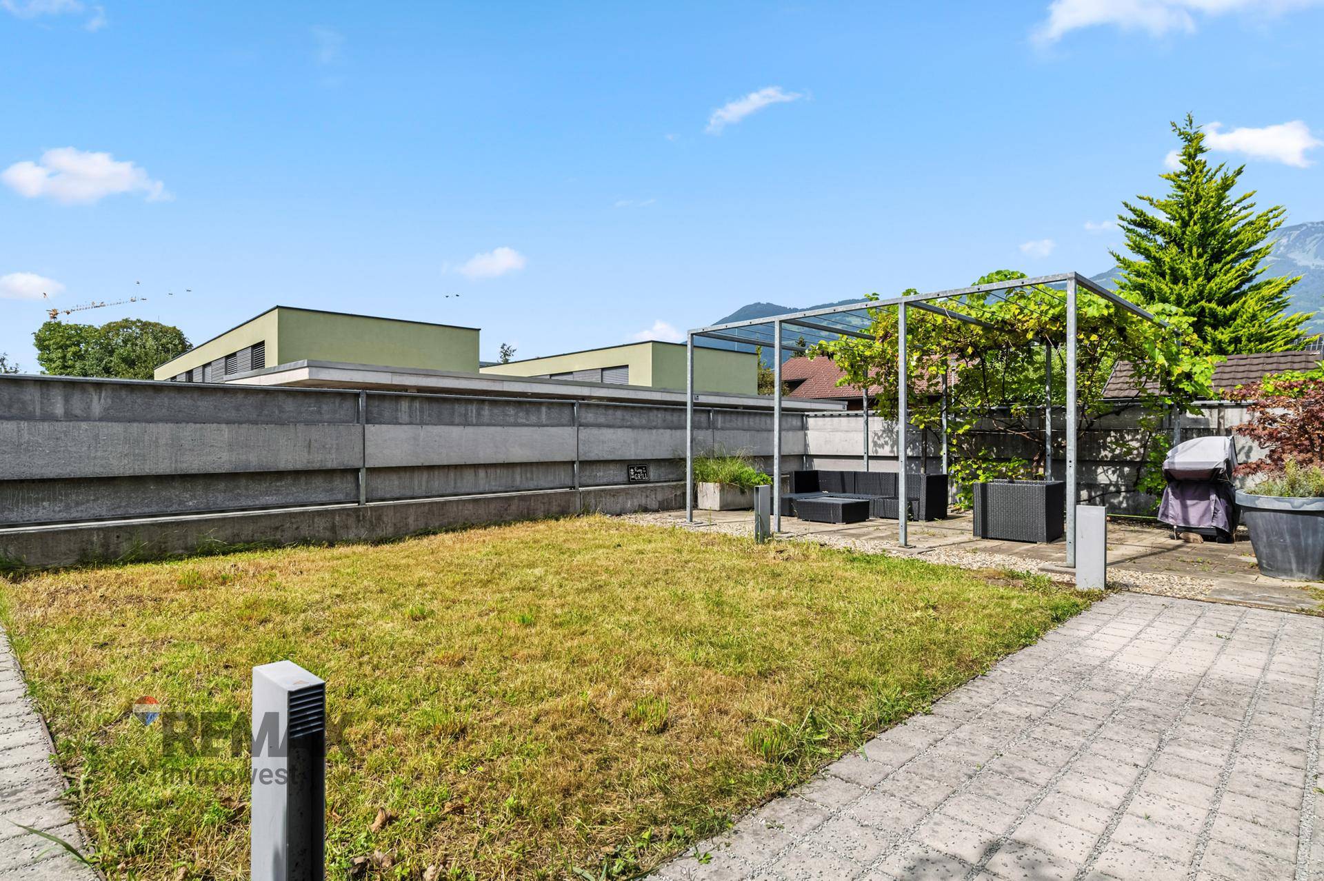 Außenbereich mit Rasenfläche, gepflastertem Weg und einer Terrasse mit Pergola im Hintergrund, umgeben von Natur.
