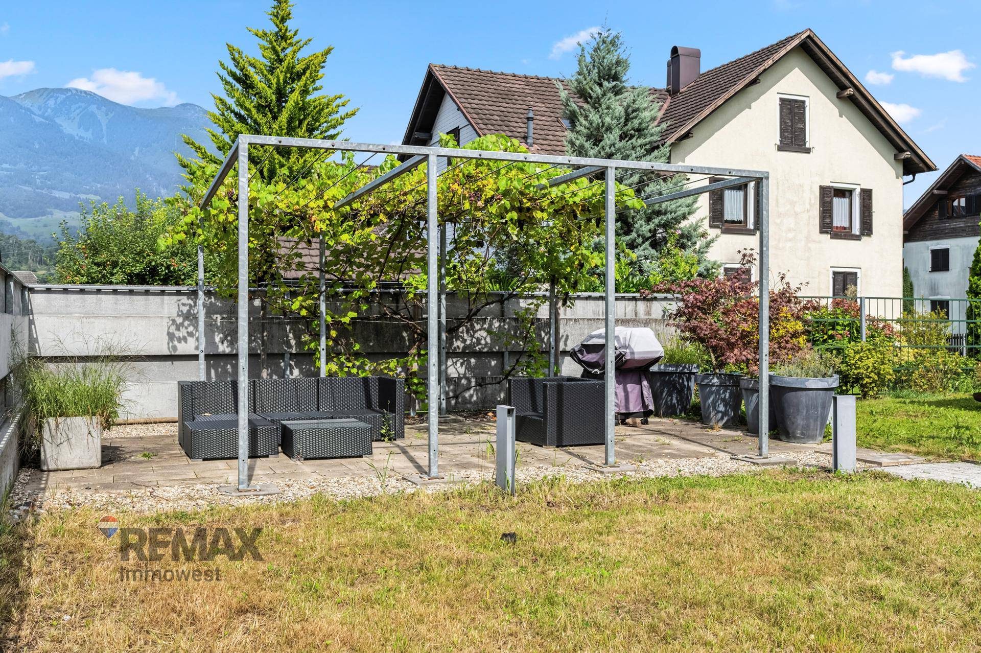 Gartenbereich mit Rasen, gepflasterter Terrasse, Pergola und Grillplatz, mit Bergblick und Wohnhaus im Hintergrund.