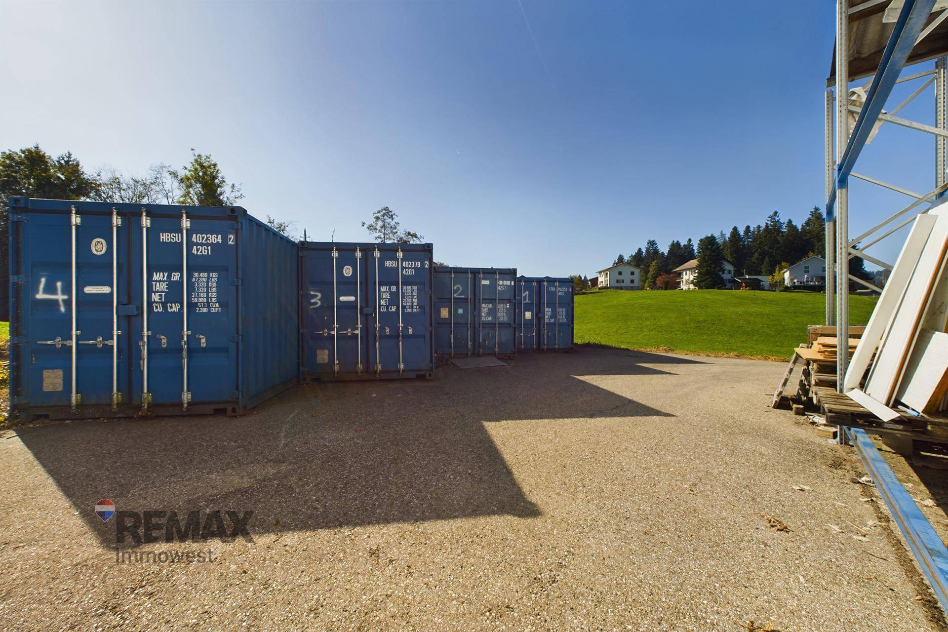 Reihe blauer Schiffscontainer auf einem Schotterplatz, umgeben von grünen Hügeln und Wohnhäusern.
