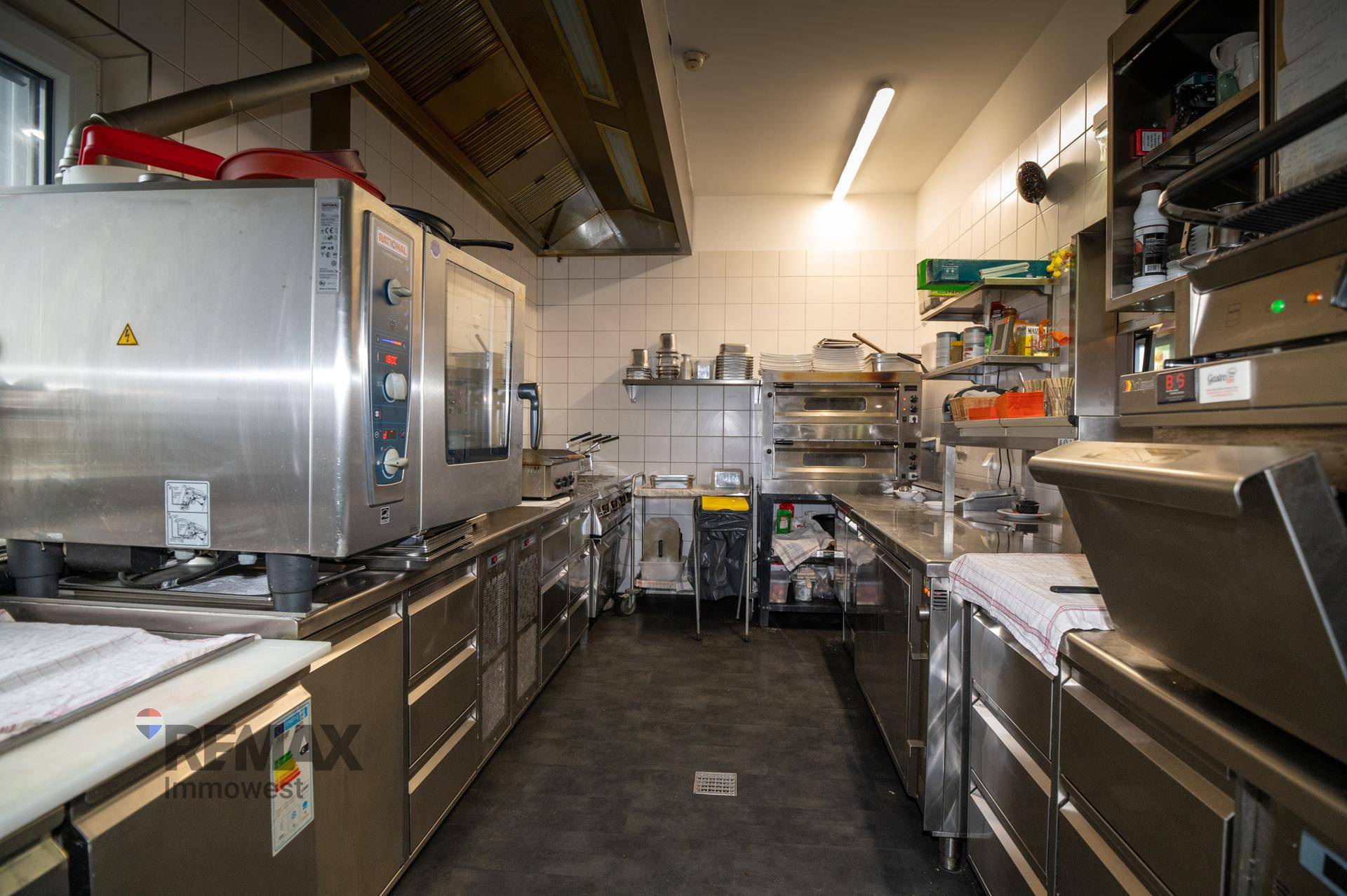 Professionelle Großküche mit Edelstahlgeräten, Abluftsystem und Fliesenwänden, sehr gut ausgestattet.