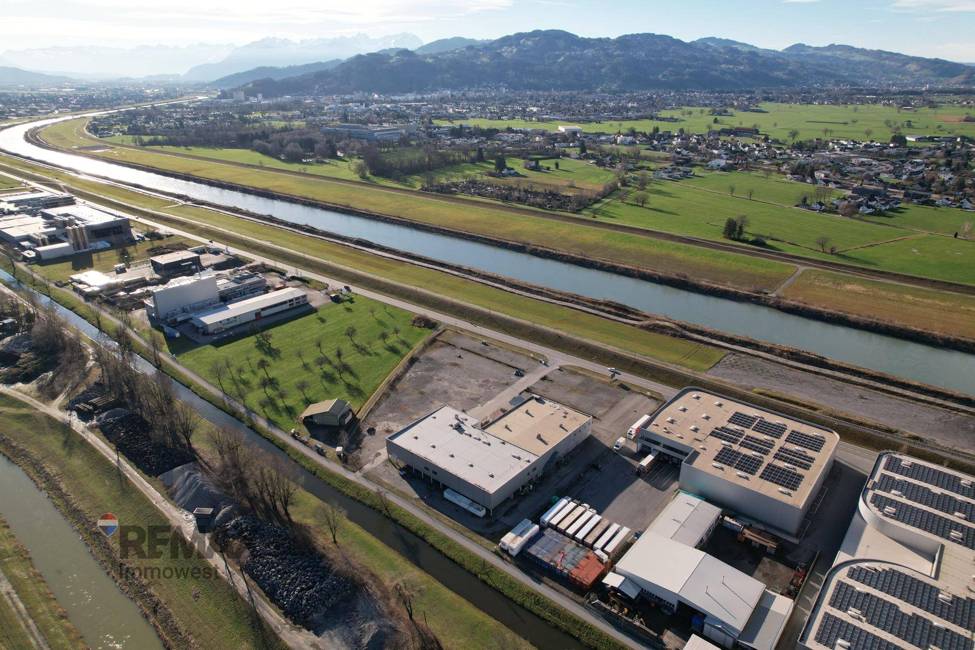 Luftaufnahme des Gewerbegebiets mit Fluss, Feldern und einer beeindruckenden Bergkulisse, malerische Umgebung.