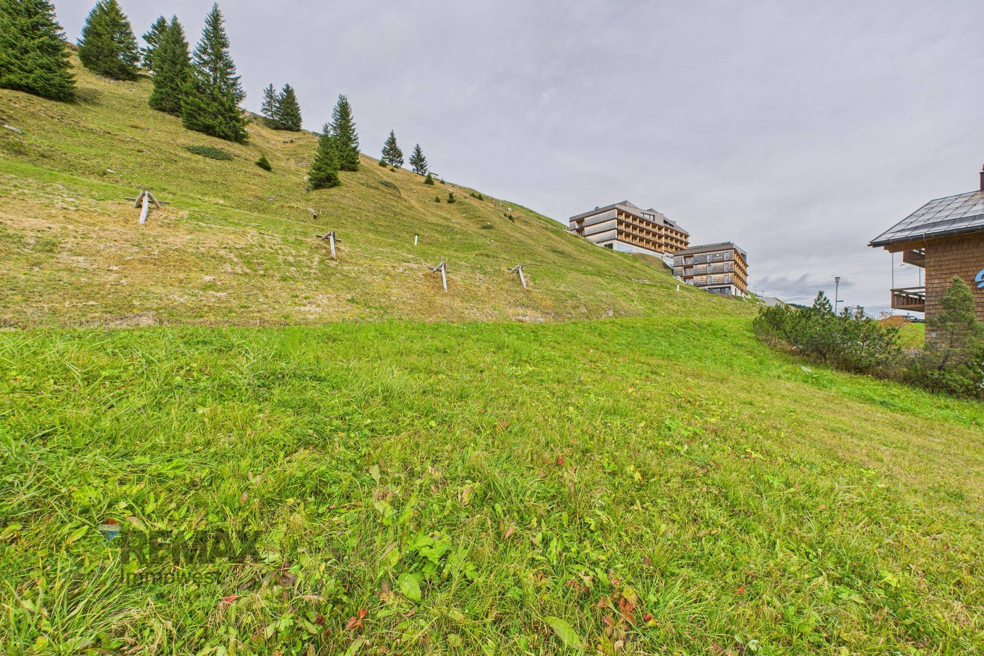 Grüne Wiese am Hang mit weiteren Gebäuden und Bergen in der Ferne unter bewölktem Himmel.
