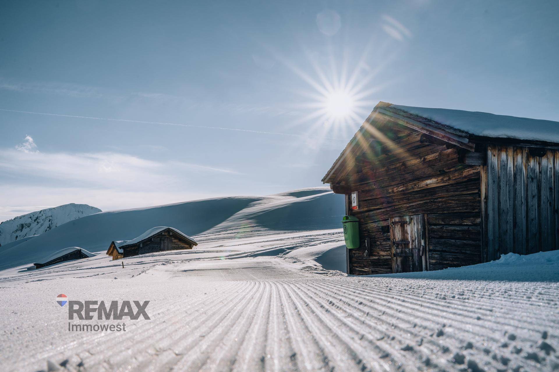 Verschneite Winterlandschaft mit traditionellen Holzhütten und präparierten Skipisten unter strahlender Sonne.