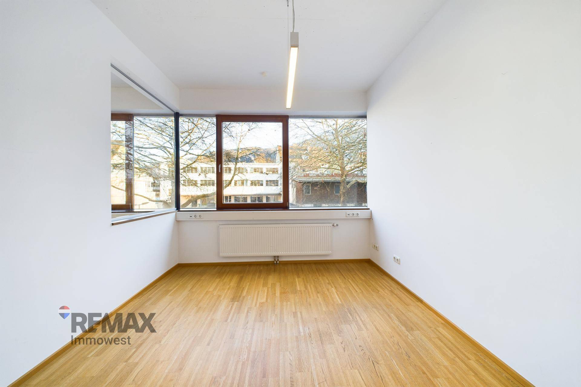 Leerer Büroraum mit Holzboden, weißen Wänden und breiten Fenstern, die viel Licht hereinlassen.
