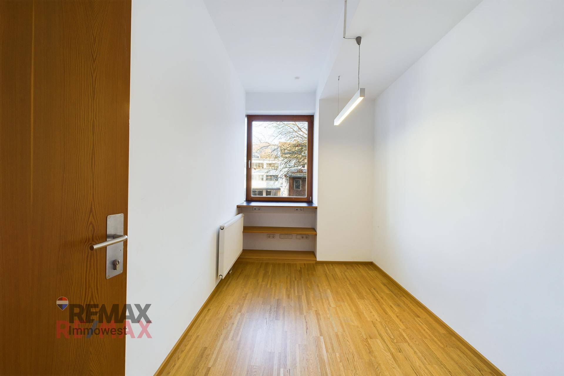 Kleiner Büroraum mit Holzboden, weißen Wänden und einem Fenster mit integrierter Ablagefläche.