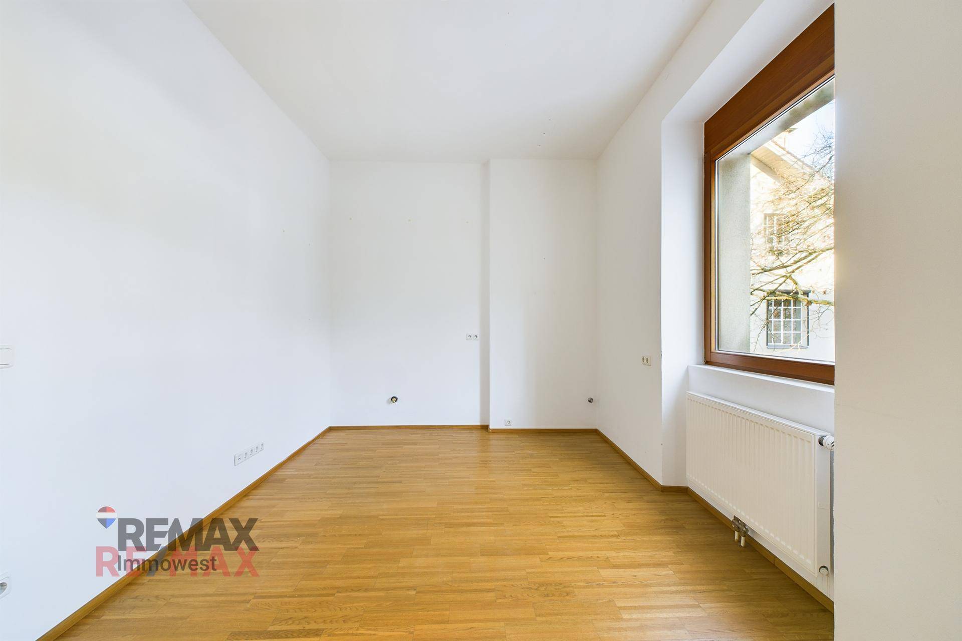Leerer Raum mit Holzboden, weißen Wänden und einem Fenster, das Tageslicht hereinlässt.