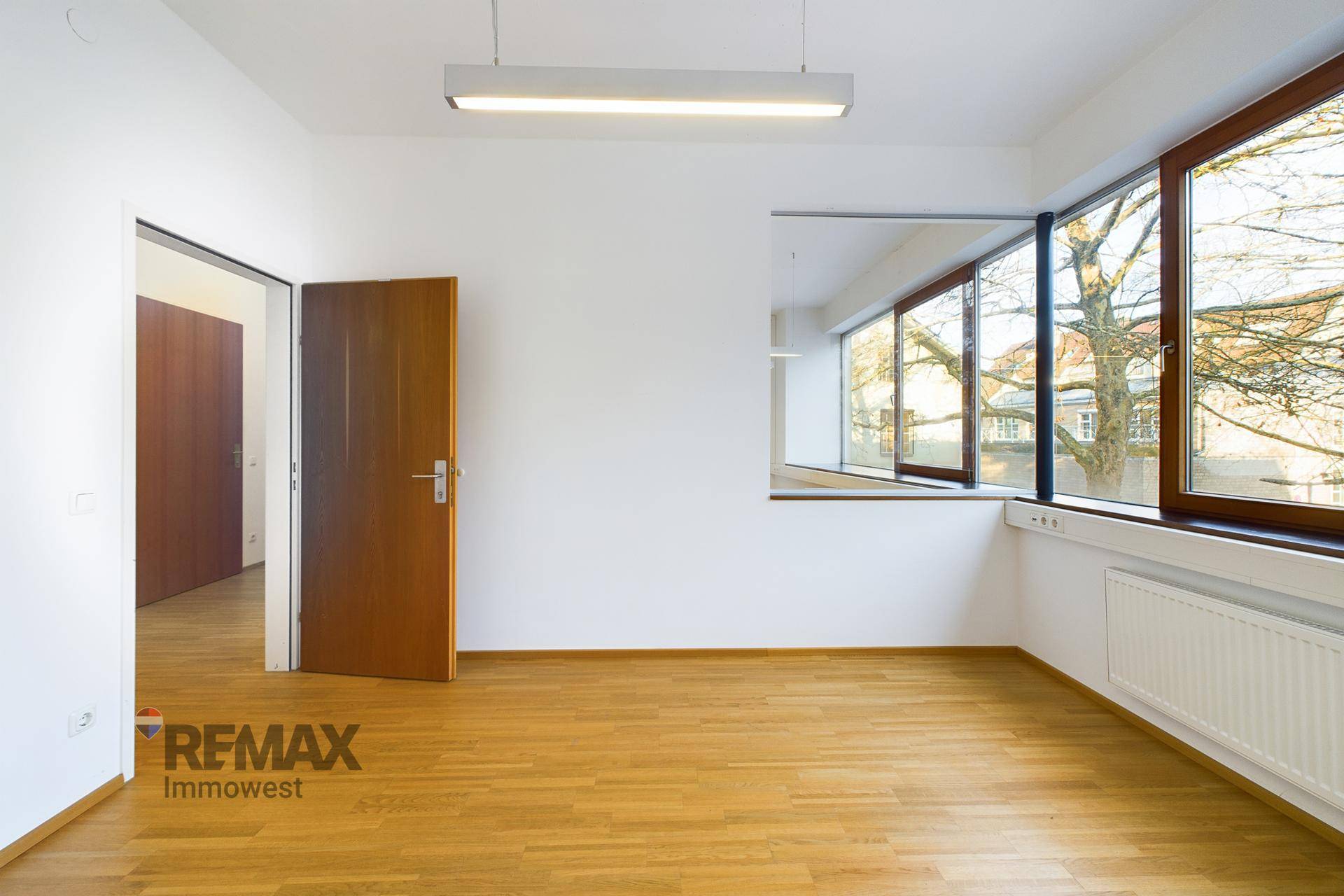 Heller Büroraum mit Holzboden, weißen Wänden und einer Holztür, die zu einem anderen Bereich führt.