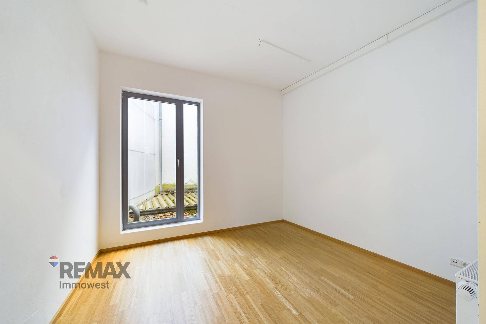 Kleinerer Büroraum mit Holzboden, weißen Wänden und einem hohen Fenster mit Blick auf einen Innenhof.