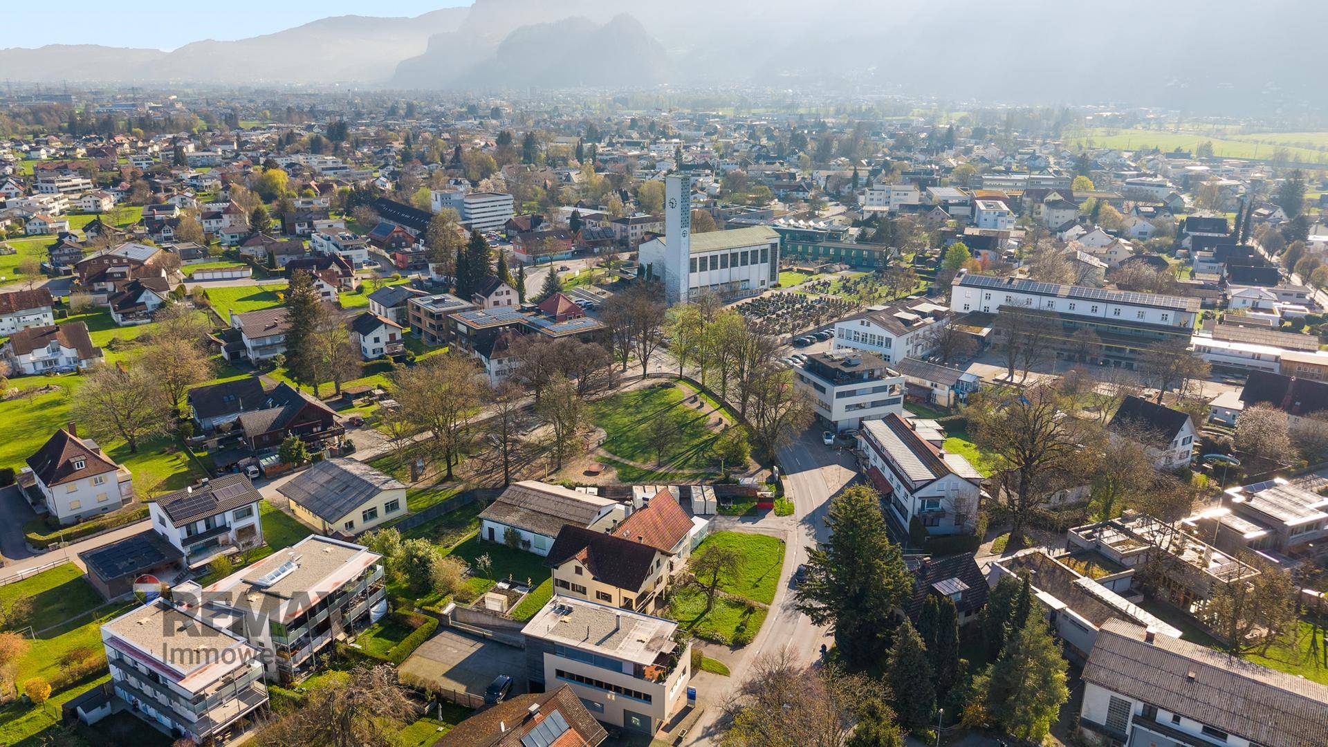 Panoramische Luftaufnahme einer Stadt mit Kirche, Wohngebäuden und Bergen am Horizont unter blauem Himmel.