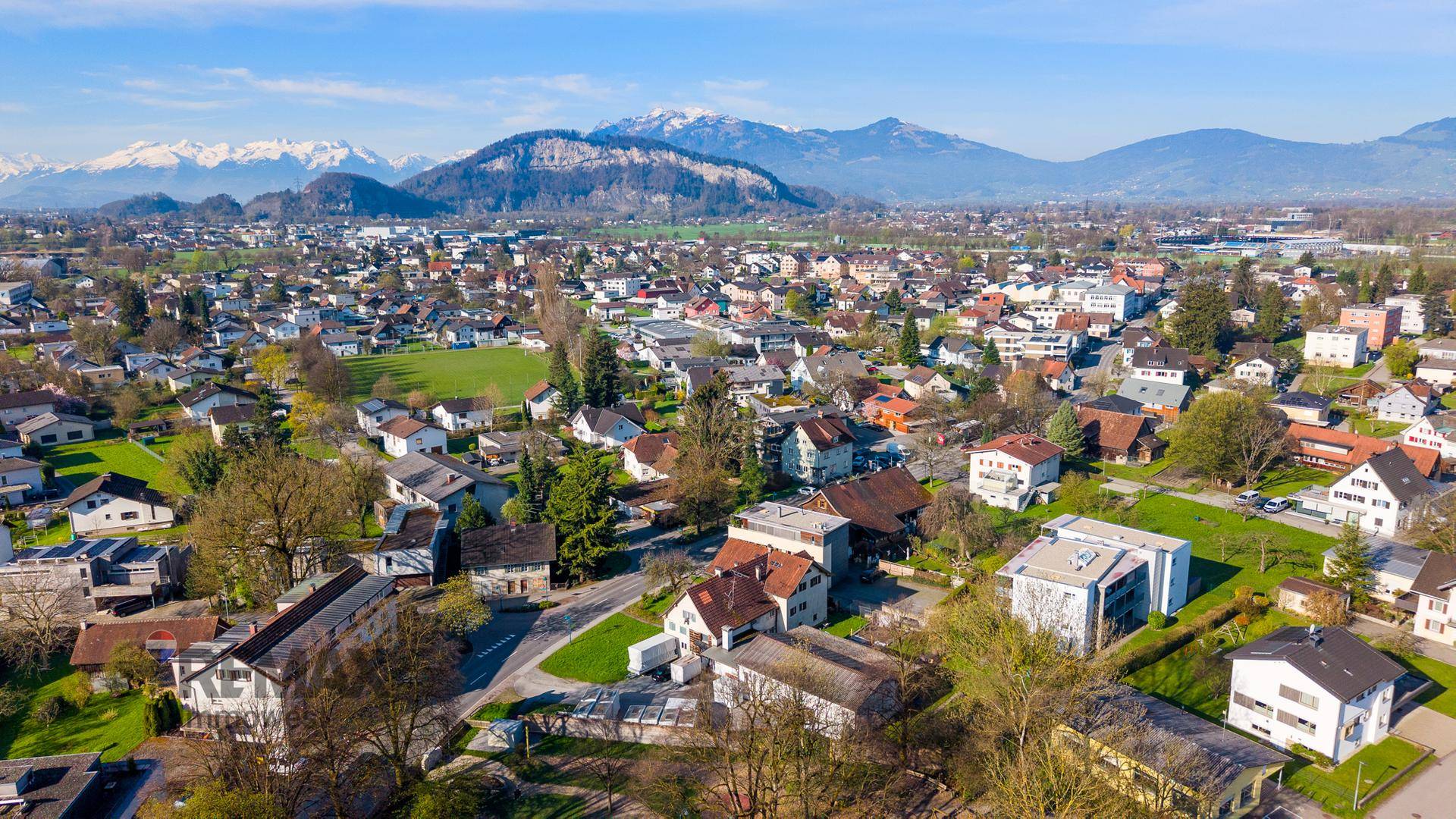 Weitläufige Luftaufnahme einer Siedlung mit vielen Häusern und schneebedeckten Bergen im Hintergrund.