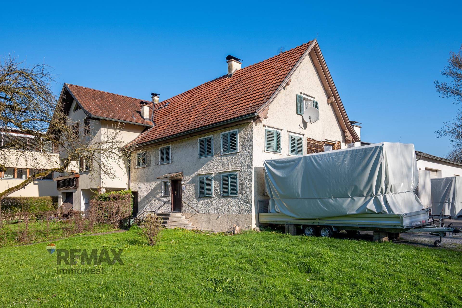 Traditionelles Wohnhaus mit hellen Fassaden, grünen Fensterläden und einem großen Vorgarten an einem sonnigen Tag.