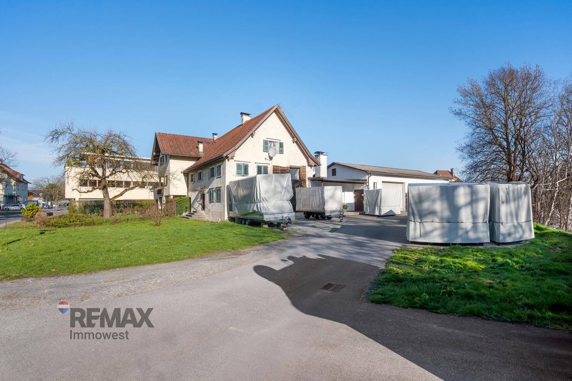 Traditionelles Wohnhaus mit großem Vorgarten und Zufahrt, umgeben von Grünflächen unter blauem Himmel.