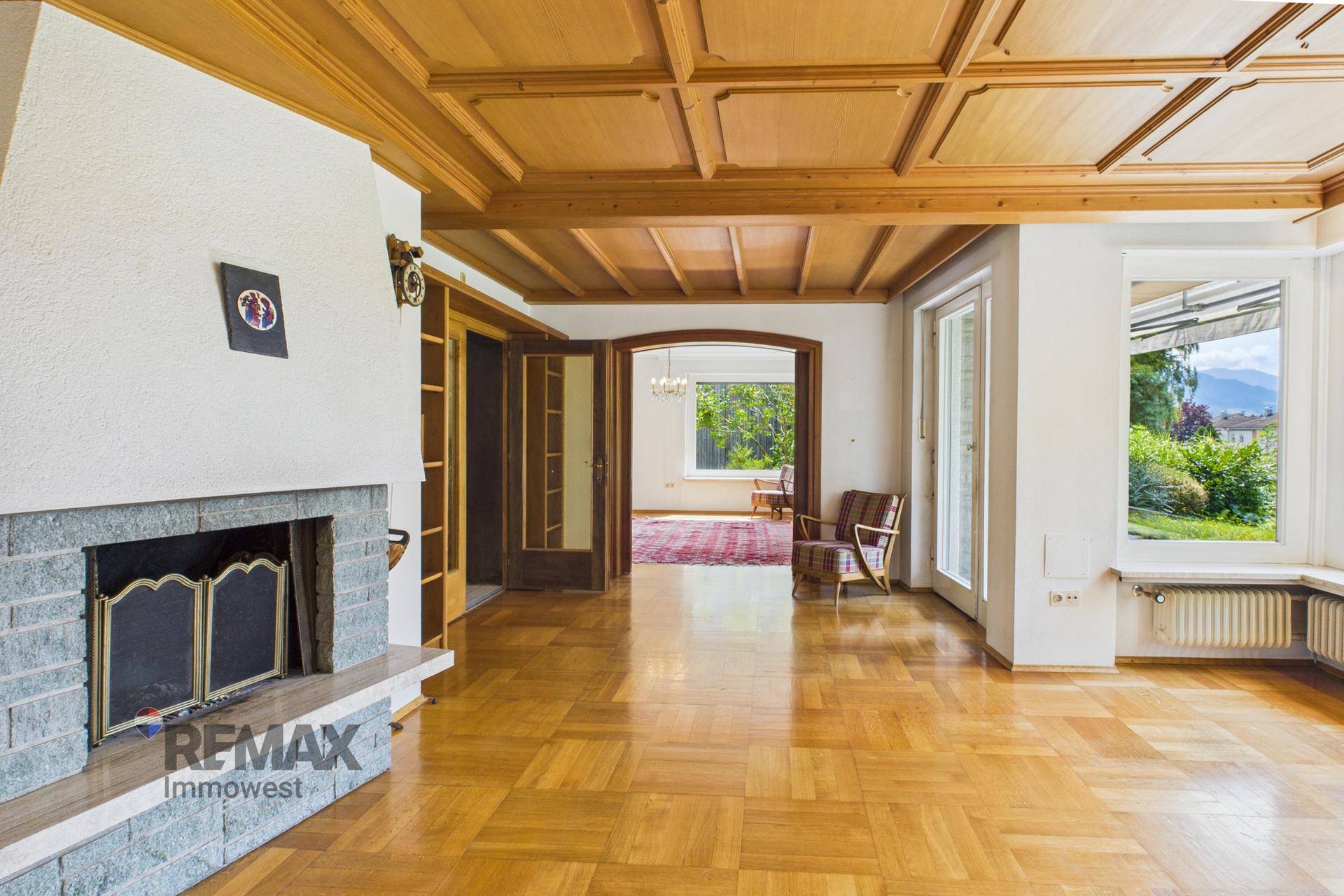 Gemütliches Wohnzimmer mit Holzkassettendecke, Parkettboden und Kamin, offen zu weiteren Räumen.