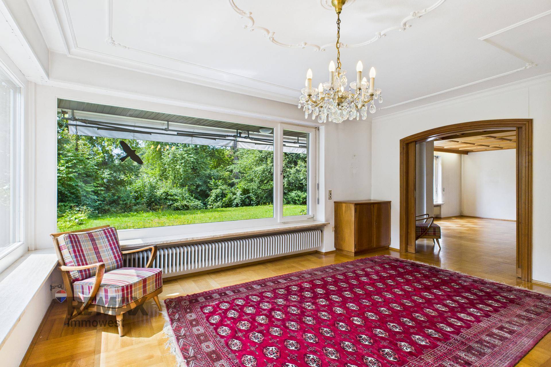 Elegantes Wohnzimmer mit Stuckdecke, Parkettboden und großem Fenster mit Blick ins Grüne.