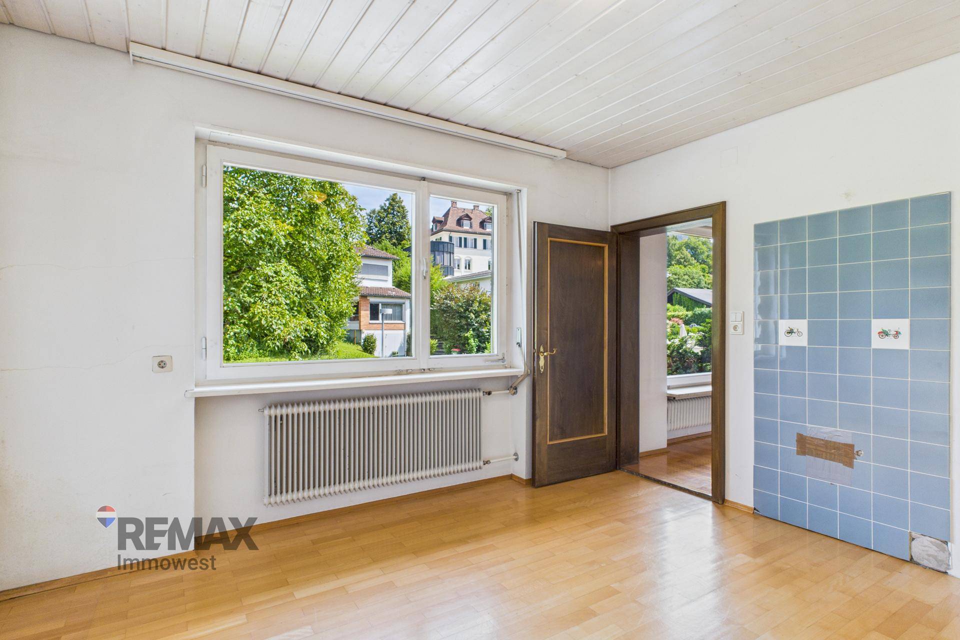 Helles Zimmer mit Parkettboden und Fenster, teilweise geflieste Wand deutet auf frühere Nutzung hin.