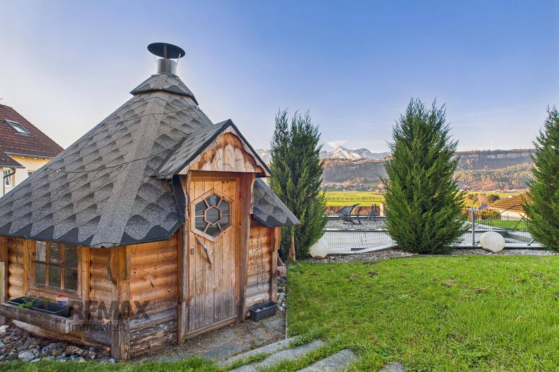 Rustikale Holzhütte im Garten mit Blick auf die Berge, umgeben von grüner Wiese und Bäumen.