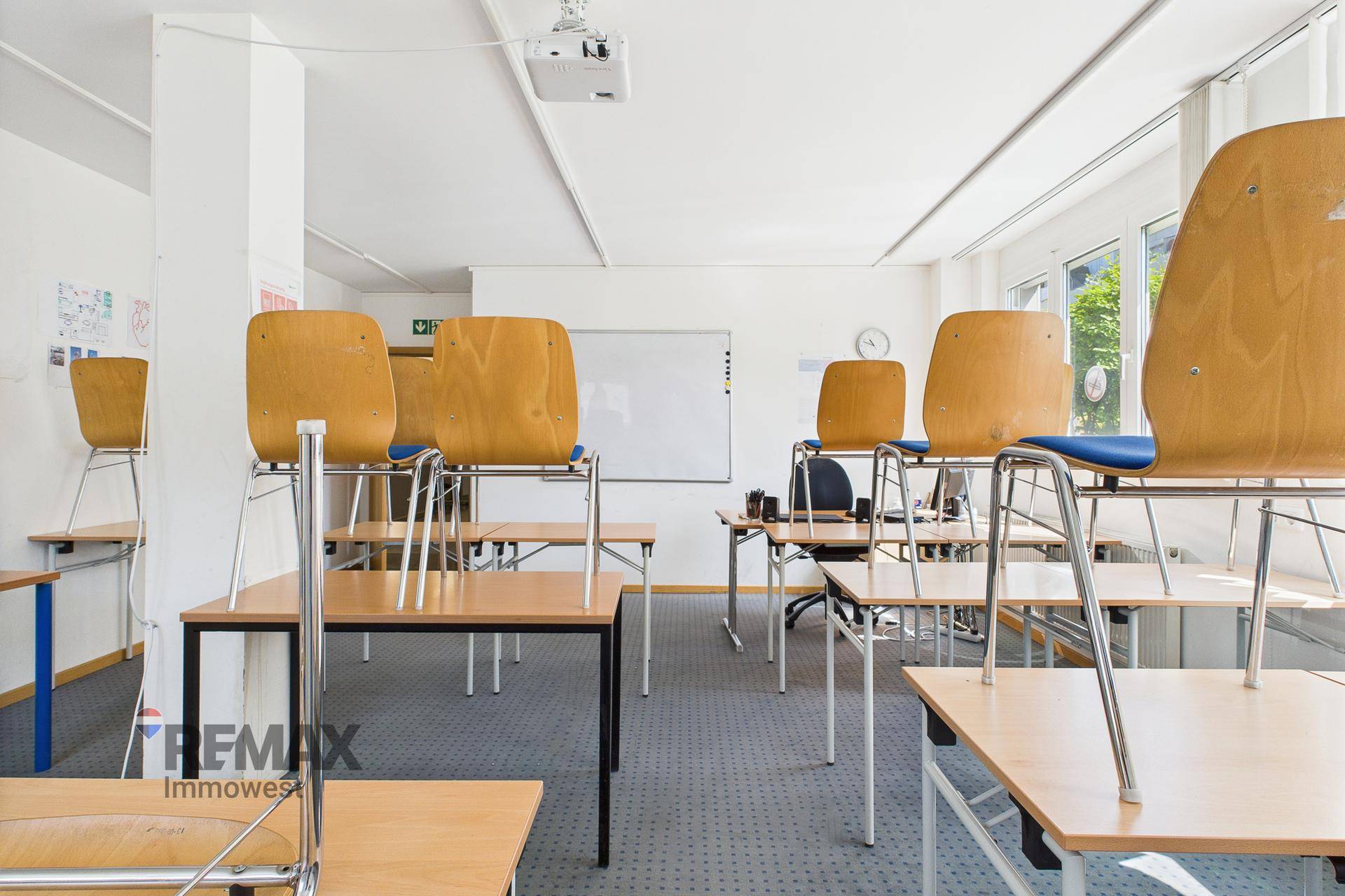 Schulungsraum mit gestapelten Stühlen und Tischen, einem Whiteboard und Teppichboden, funktional eingerichtet.