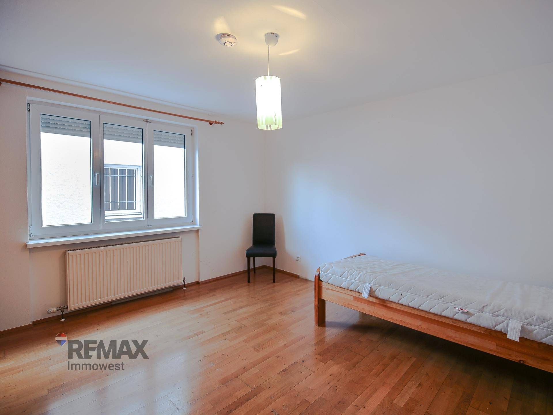 Helles Schlafzimmer mit Holzboden, großem Fenster und einfacher Einrichtung.