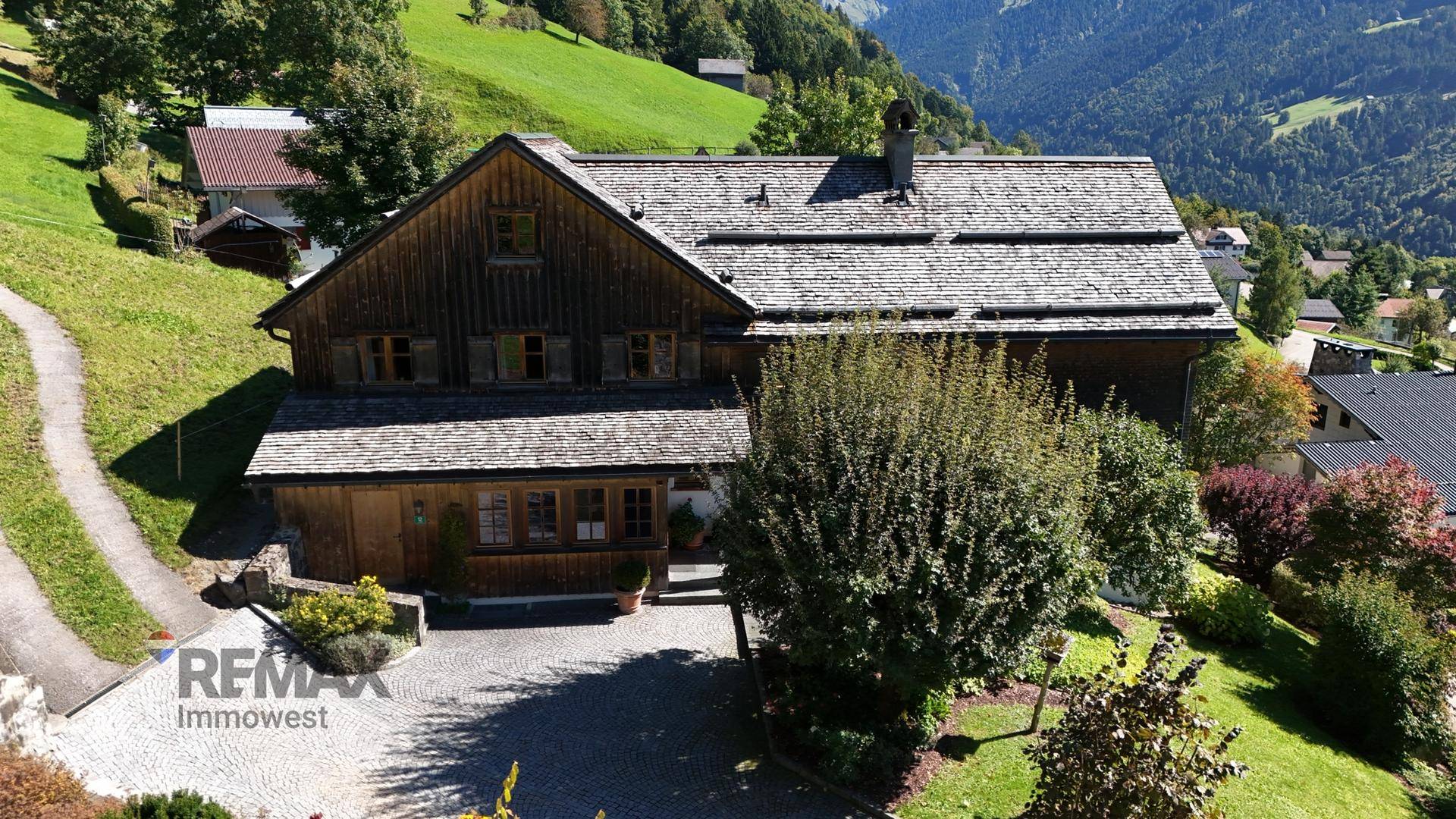 Traditionelles Holzhaus mit Schindeldach, umgeben von grünen Hügeln und Bergen in ländlicher Umgebung.